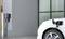 Ein weißes Elektroauto wird an einer Ladestation aufgeladen. Das Fahrzeug ist vor einem modernen Gebäude geparkt.