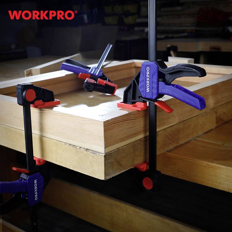 Eine Holzplatte wird mit mehreren Zwingen der Marke WorkPro an einem Tisch befestigt. Die Umgebung scheint eine Werkstatt zu sein.