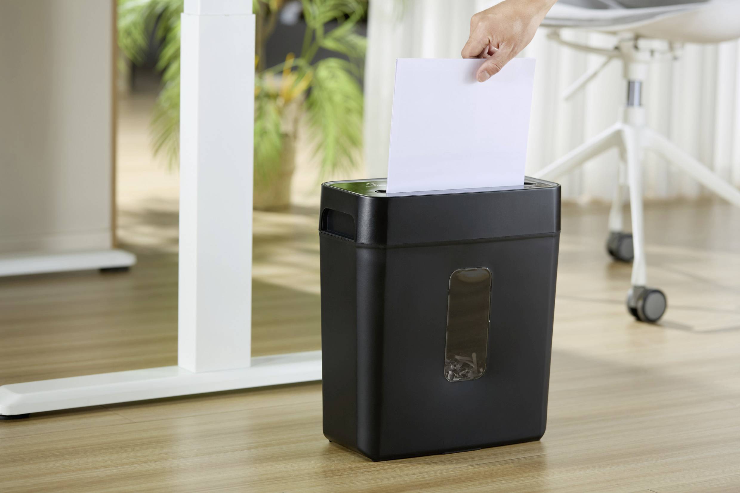 Eine Person legt ein Blatt Papier in einen schwarzen Aktenvernichter, der neben einem weißen Tisch in einem Büro steht.