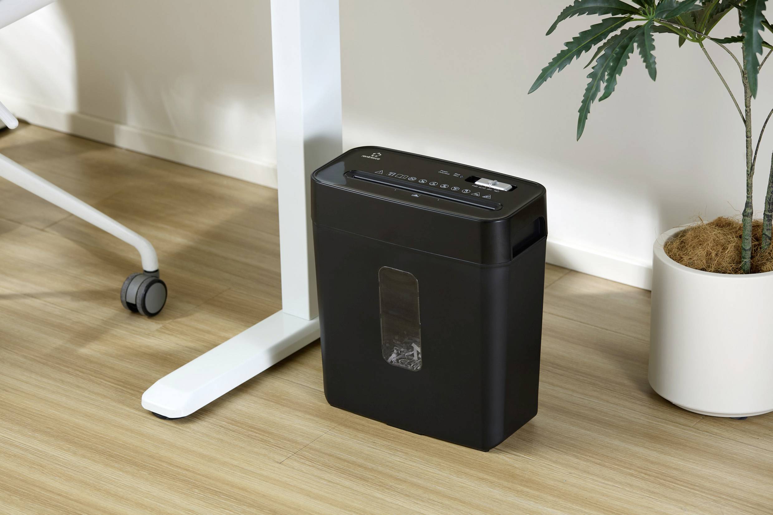 Ein schwarzer Aktenvernichter steht neben einem weißen Tischbein auf einem Holzboden. Daneben befindet sich eine grüne Pflanze im Topf.