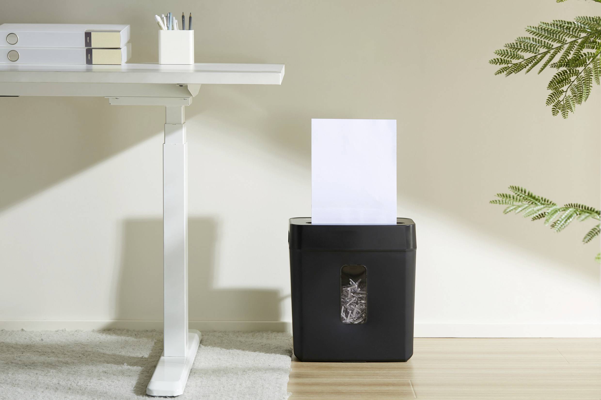 Auf einem hellen Teppich steht ein Aktenvernichter unter einem weißen Tisch; ein Blatt Papier wird gerade geschreddert.