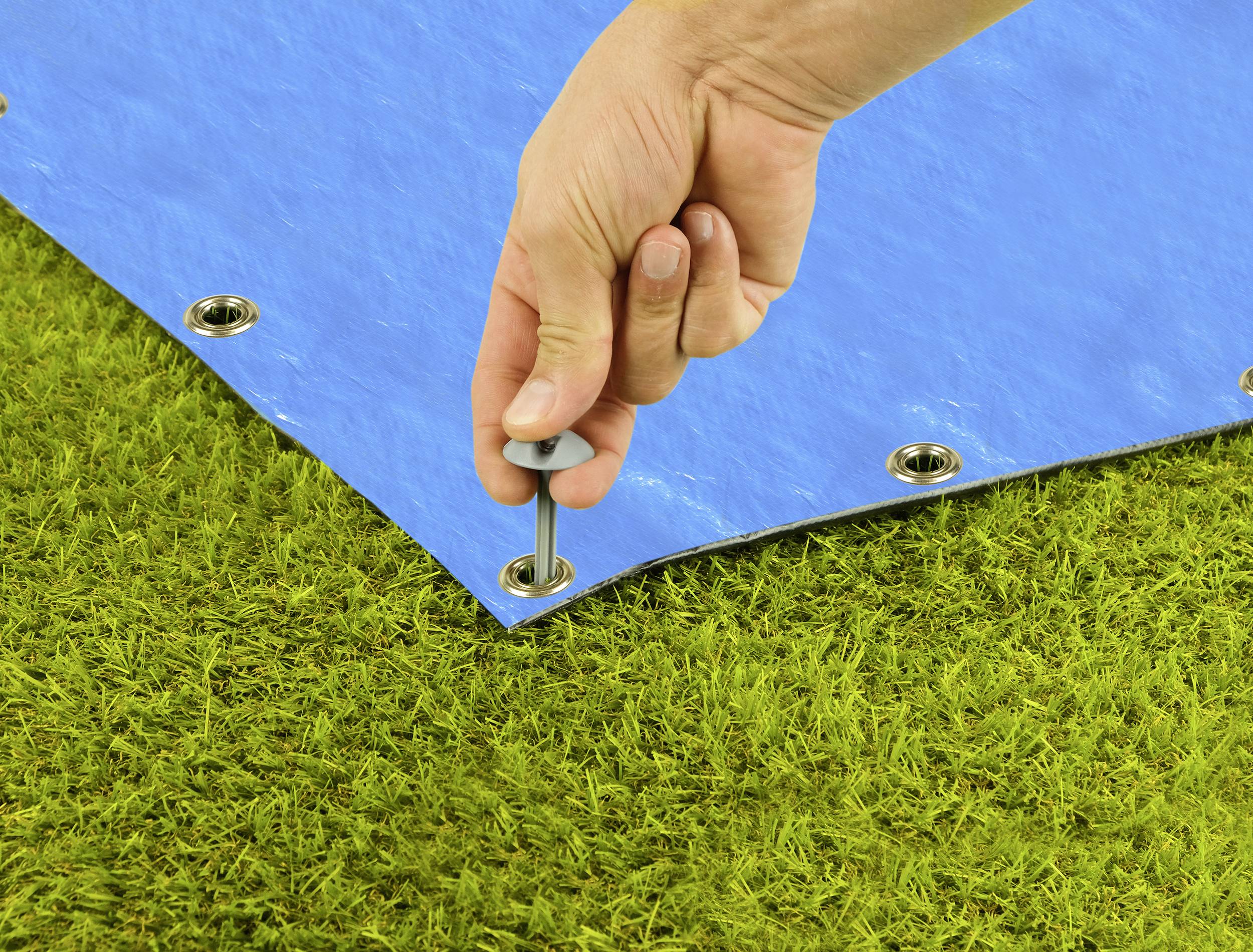 Eine Hand befestigt eine blaue Plane mit einem Bolzen auf grünem Gras. Die Plane hat Ösen zur Fixierung.
