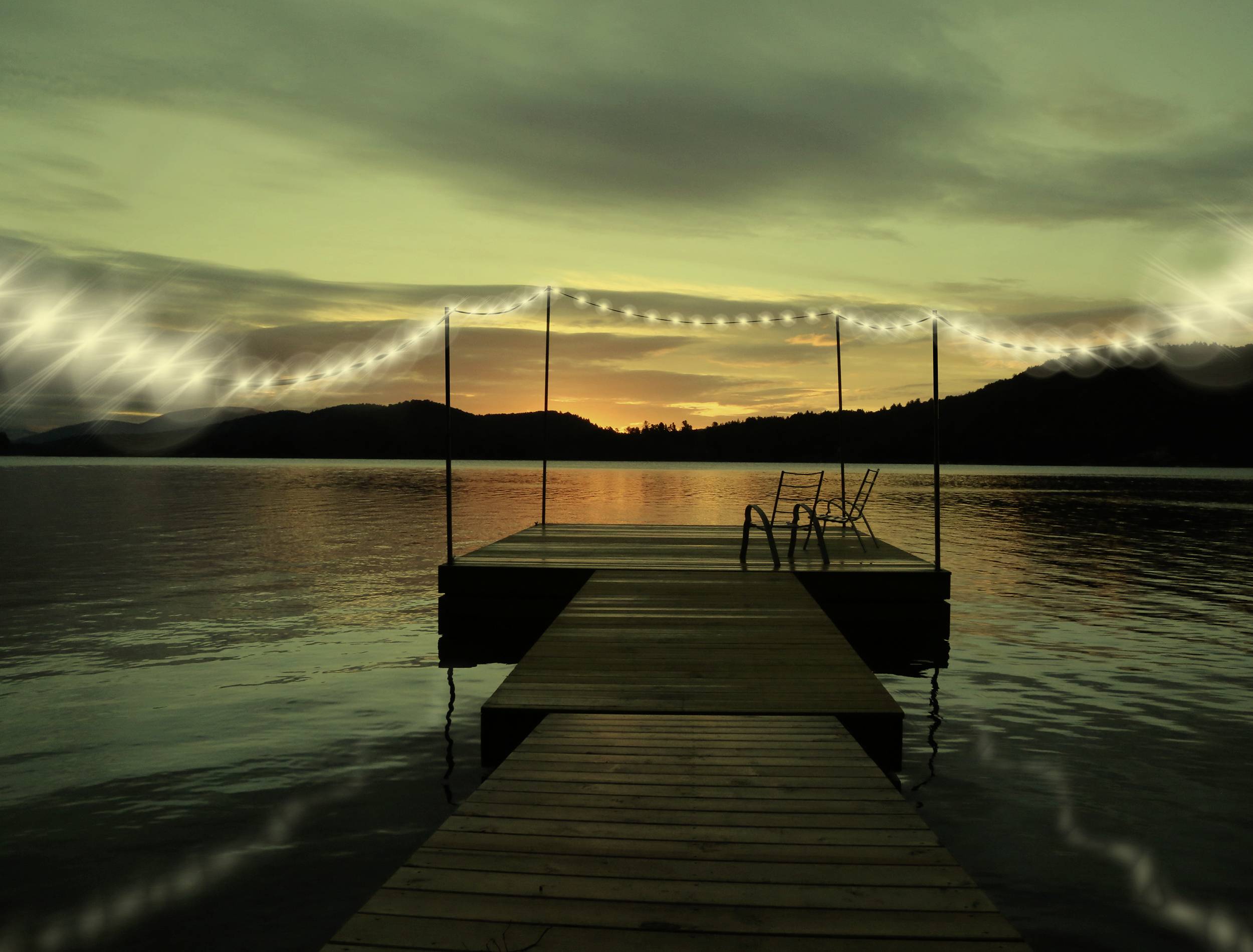 Ein Steg führt auf einen See bei Sonnenuntergang. Lichterketten hängen am Rand. Berge und dunkler Himmel im Hintergrund.