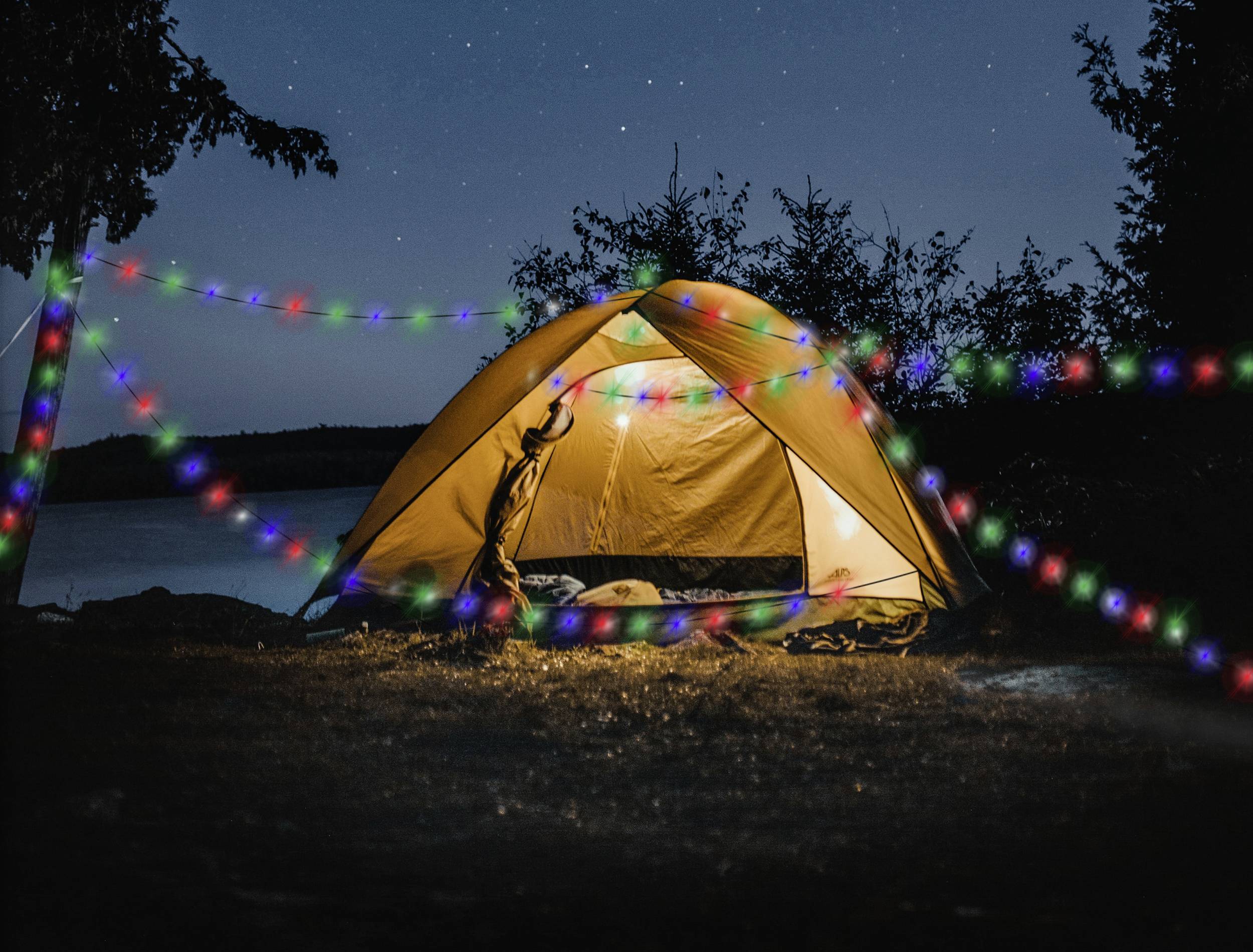 Ein gelbes Zelt steht nachts beleuchtet an einem Seeufer, umgeben von bunten Lichterketten und Sternen am Himmel.