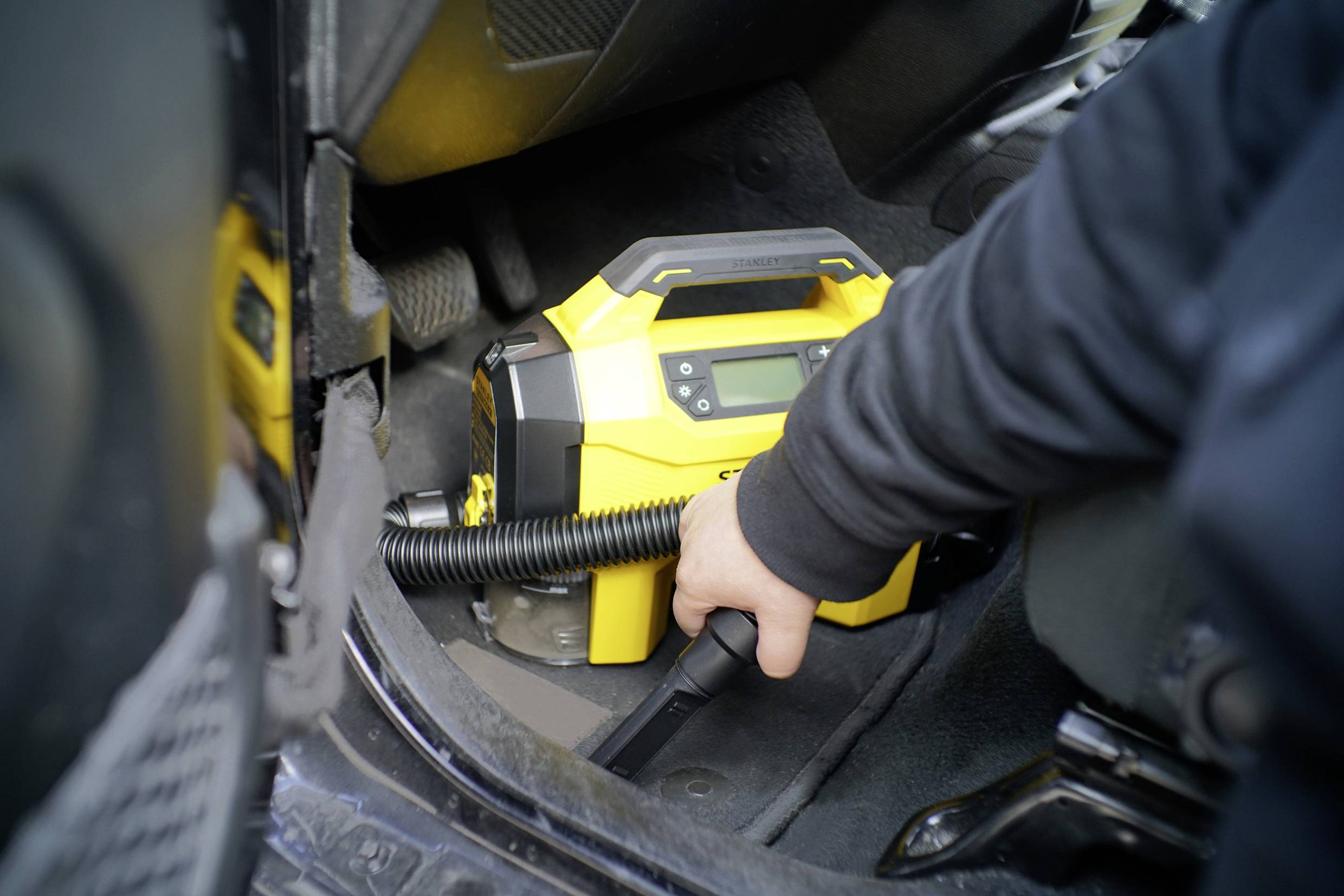 Eine Person benutzt einen gelben, tragbaren Staubsauger im Fußraum eines Autos, um den Innenraum zu reinigen.