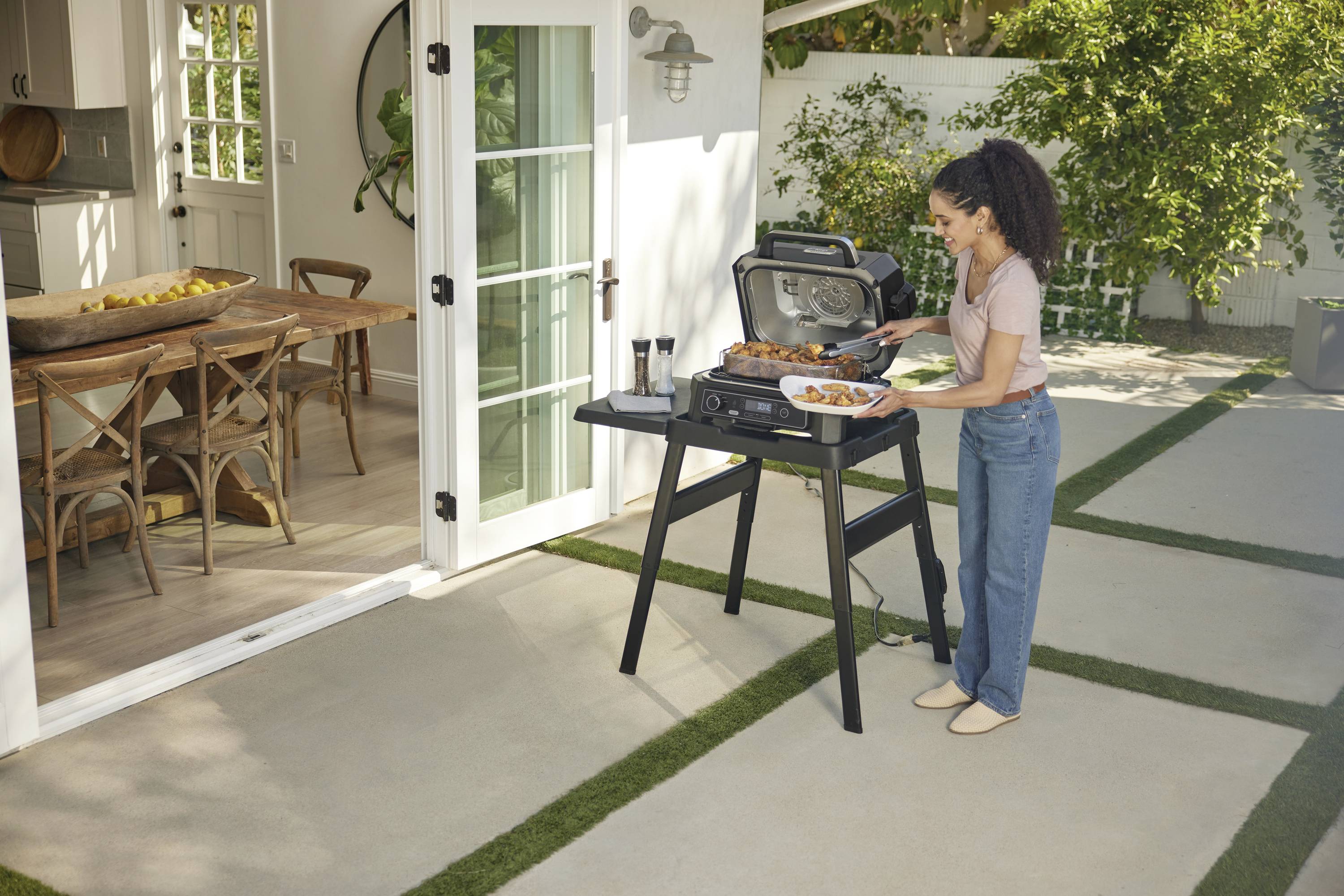 Eine Person grillt im Garten mit einem Gasgrill. Neben den Glastüren steht ein Tisch mit Stühlen im Innenbereich.