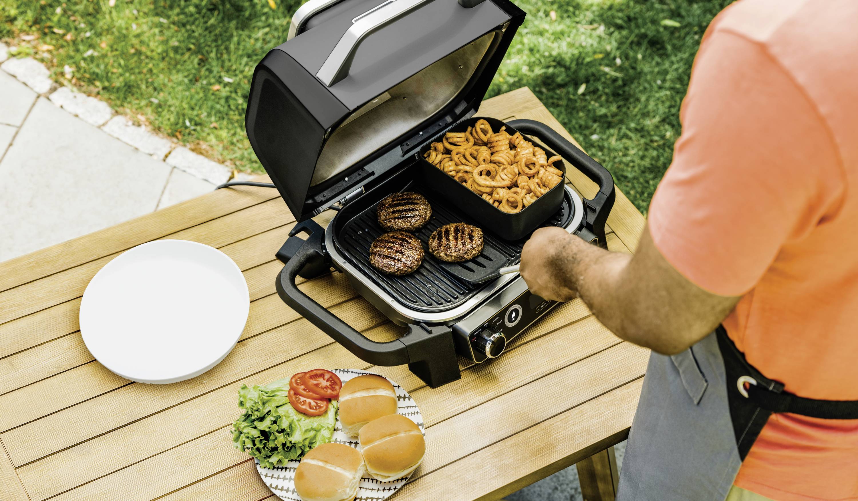 Person grillt Burger-Patties und Zwiebelringe im Freien auf einem Tisch. Teller mit Brötchen, Tomaten und Salat neben dem Grill.