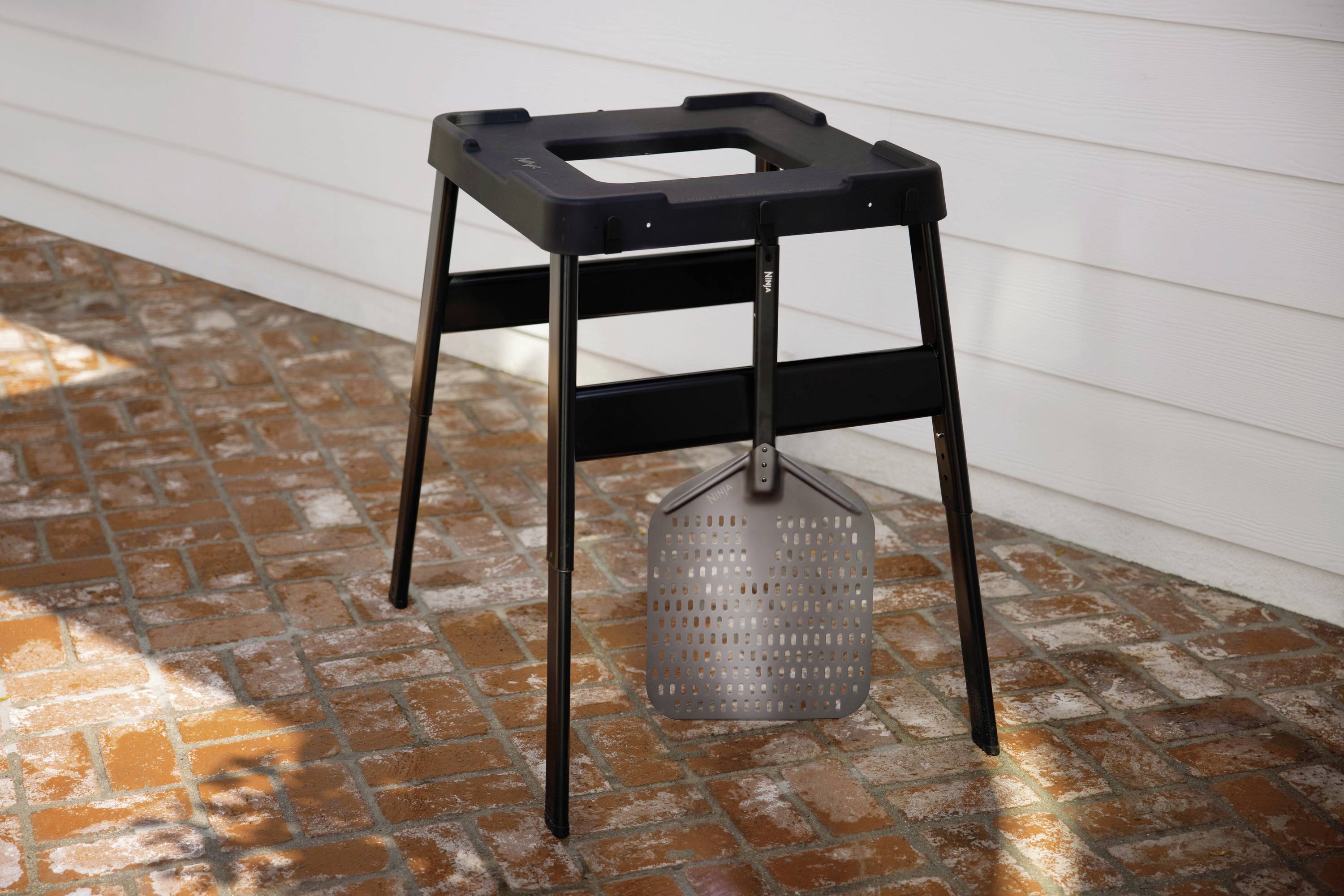 Ein metallener Outdoor-Pizzaständer steht auf einem Terrassenboden aus roten Ziegelsteinen. Am Gestell hängt eine Pizzaschaufel.