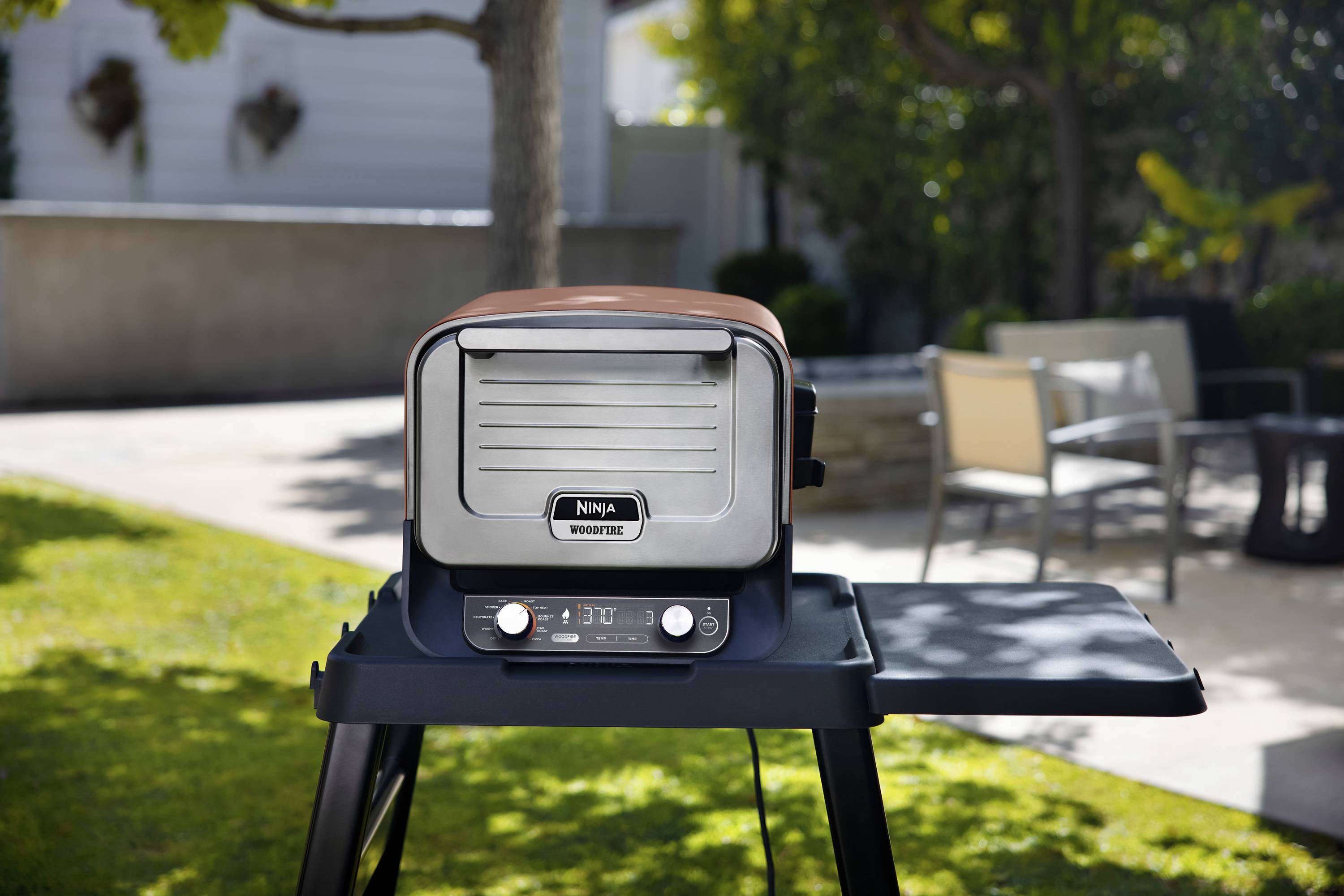 Ein elektrischer Grill steht auf einer Terrasse im Garten. Sonne scheint, umgeben von Gartenmöbeln und Bäumen.