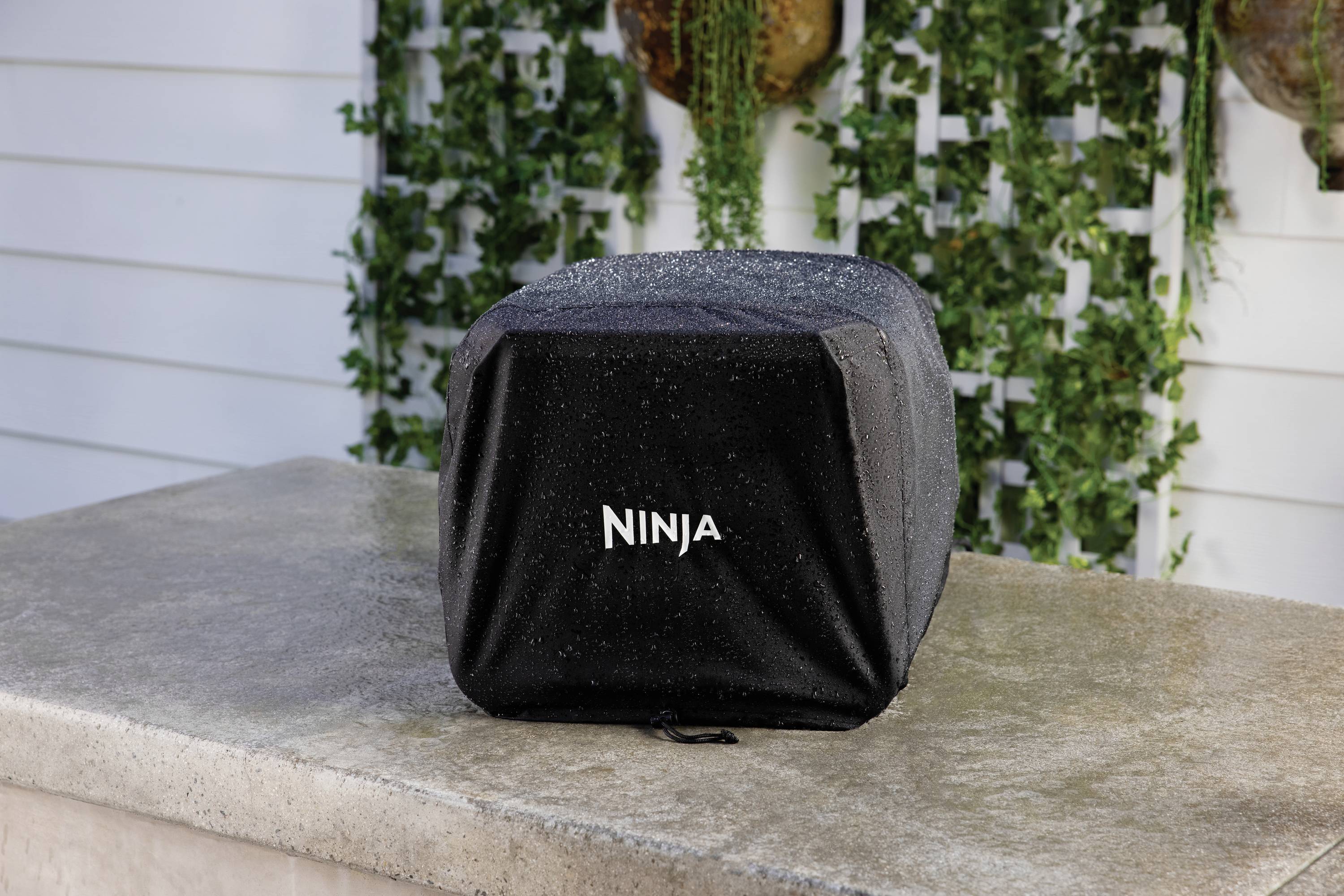 Ein schwarzer Bezug mit der Aufschrift 'NINJA' bedeckt ein Gerät auf einem steinernen Tisch. Im Hintergrund sind Pflanzen sichtbar.