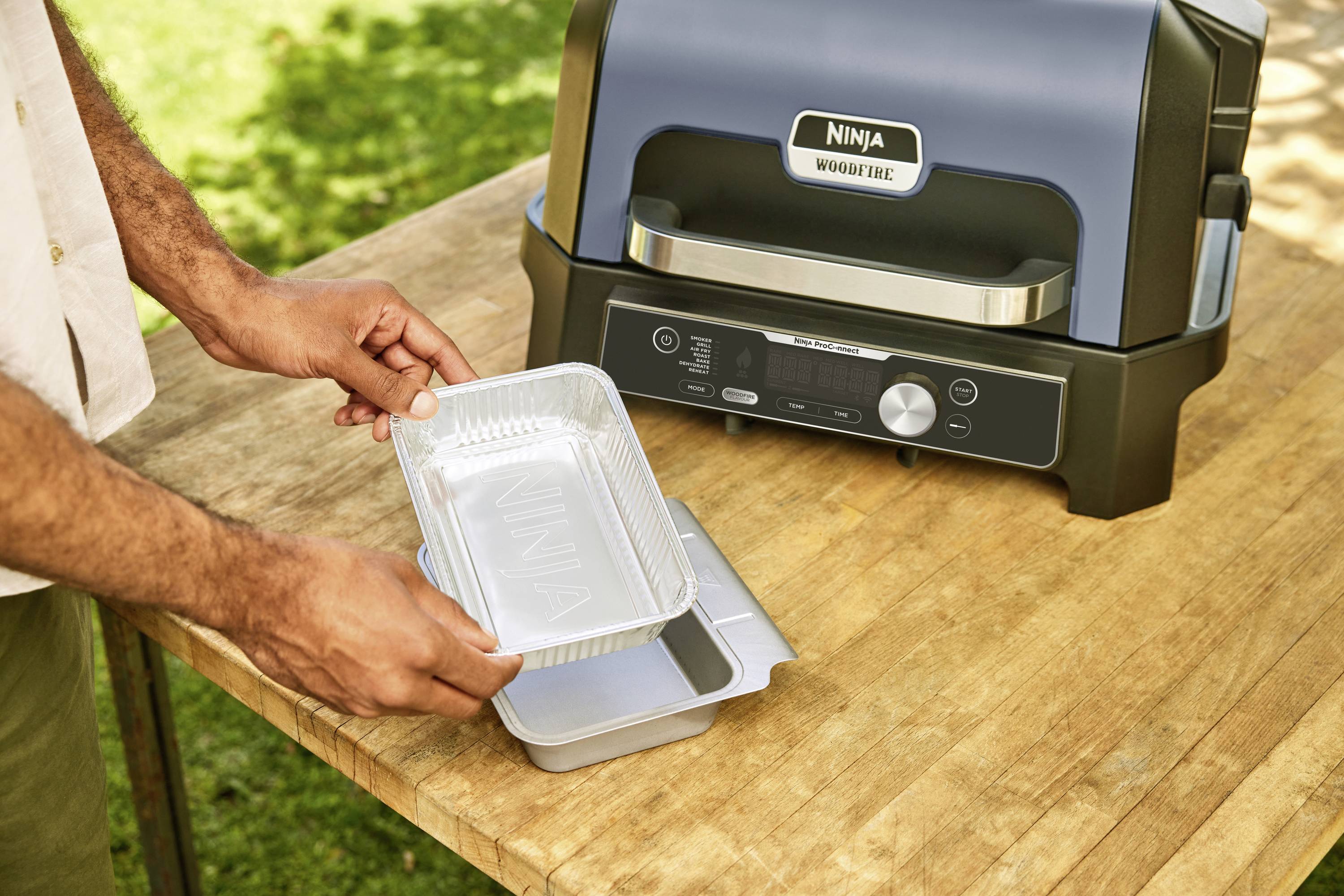 Eine Person legt eine Aluschale auf einen Grilltisch neben einem elektrischen Grill.