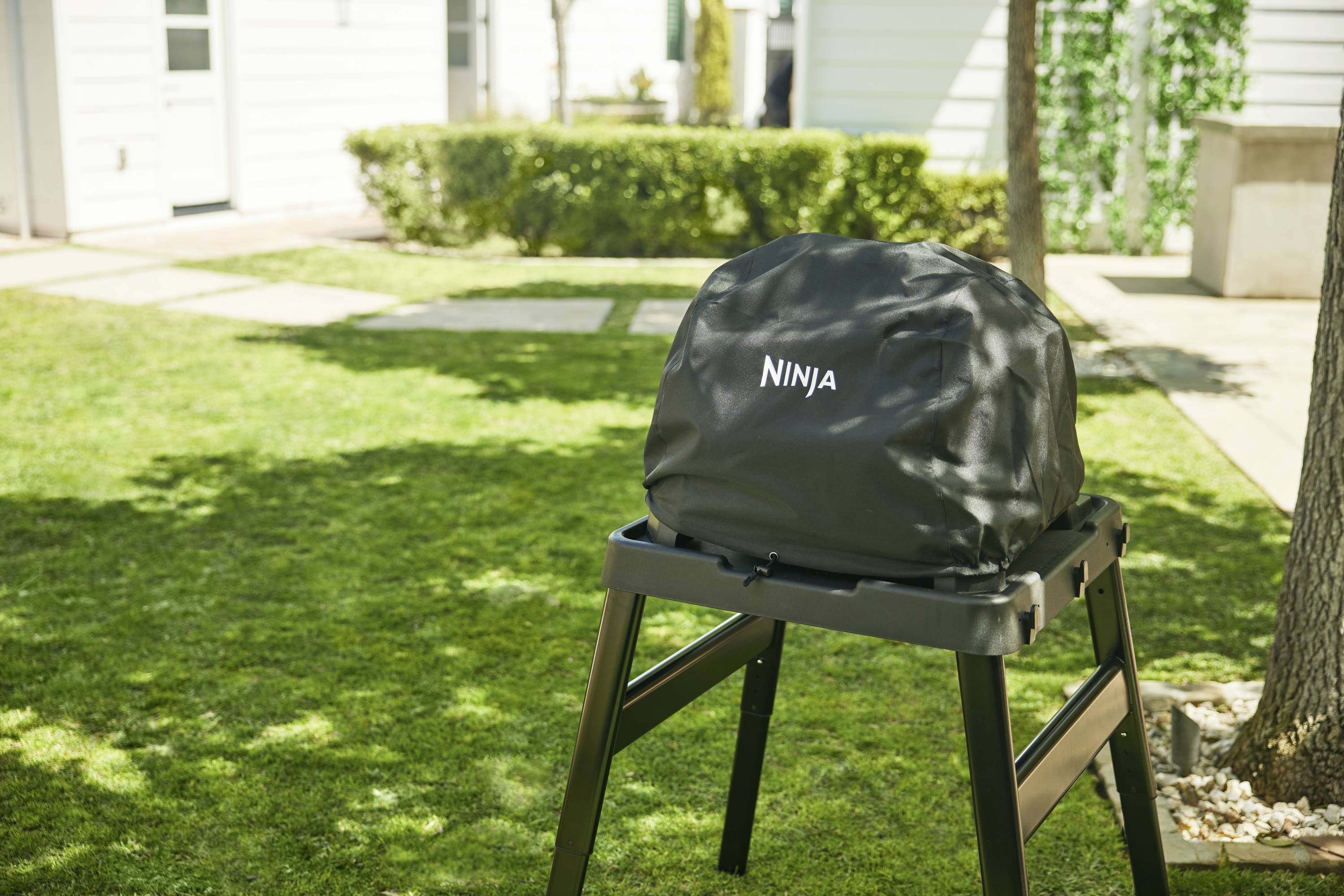 Grill im Garten mit einer schwarzen Abdeckung, auf der 'Ninja' steht. Umgebung mit Rasen, Gehweg und weißen Gebäuden im Hintergrund.