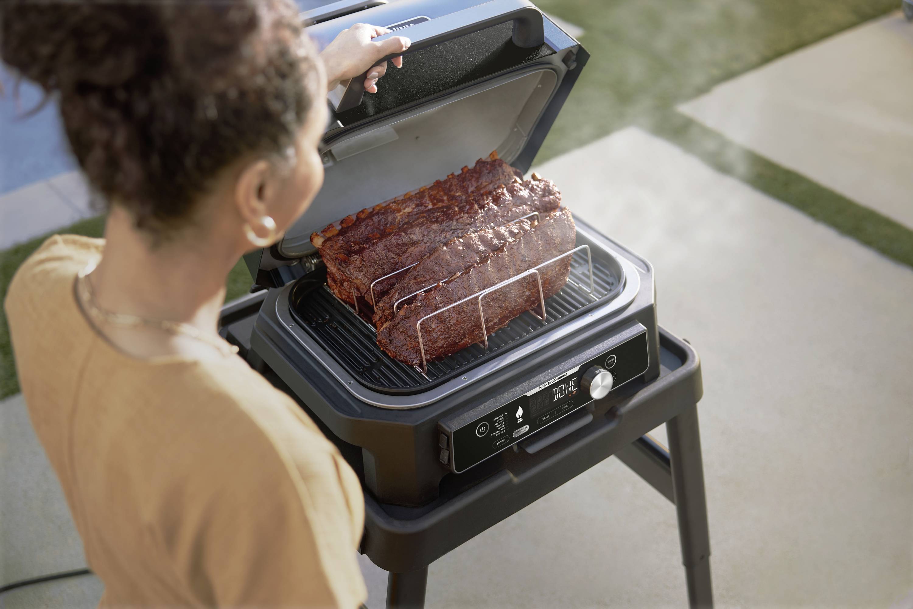 Eine Person grillt Rippchen auf einem Elektrogrill im Freien bei Tageslicht. Der Deckel des Grills ist zur Kontrolle geöffnet.