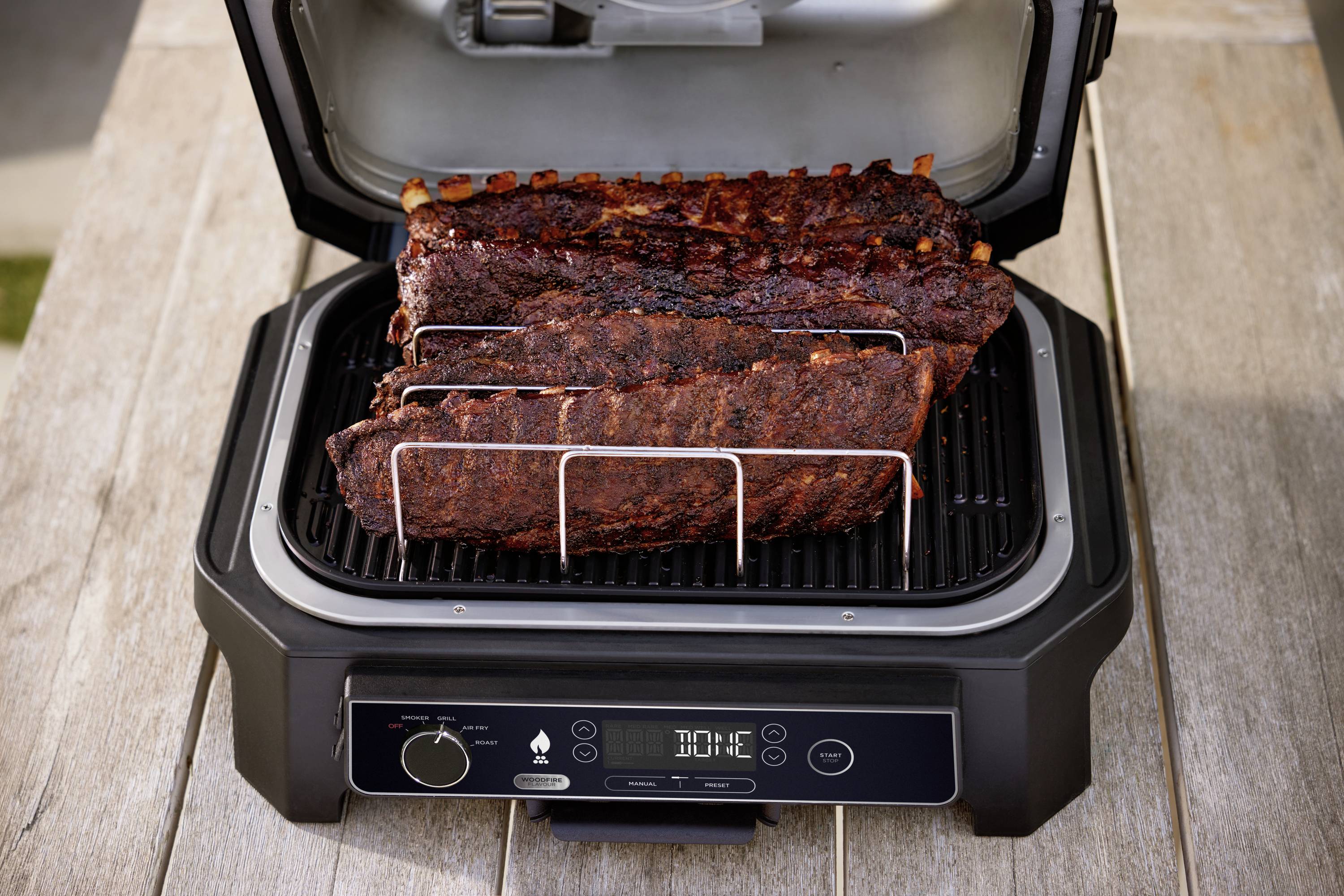 Ein elektrischer Grill auf einem Holztisch mit geöffnetem Deckel. Darin liegen drei große, dunkle, gegrillte Fleischstücke auf einem Rost.