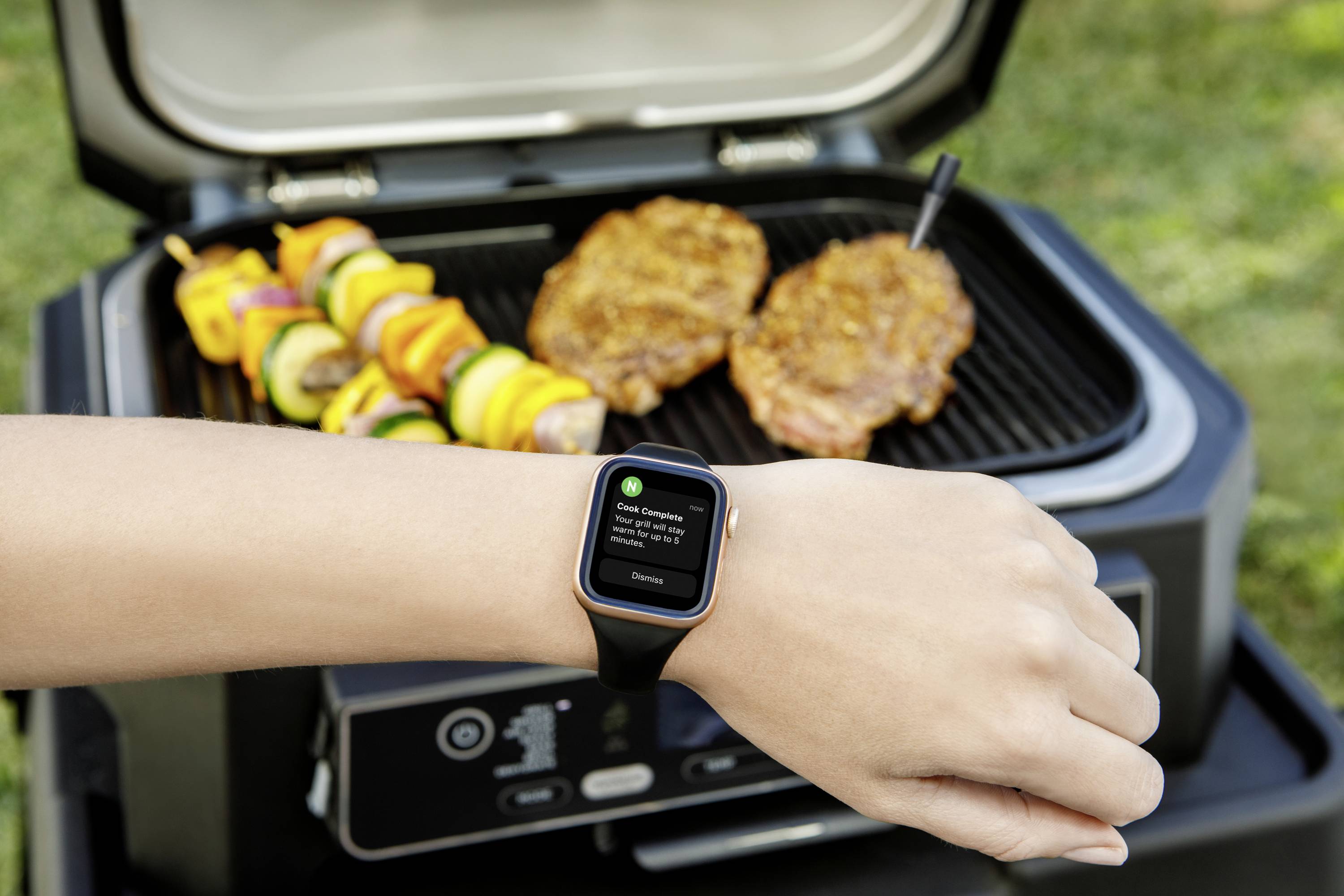 Eine Hand mit Smartwatch zeigt Nachricht: 'Fleisch umdrehen' vor einem Grill mit Fleisch und Gemüse auf Spießen.