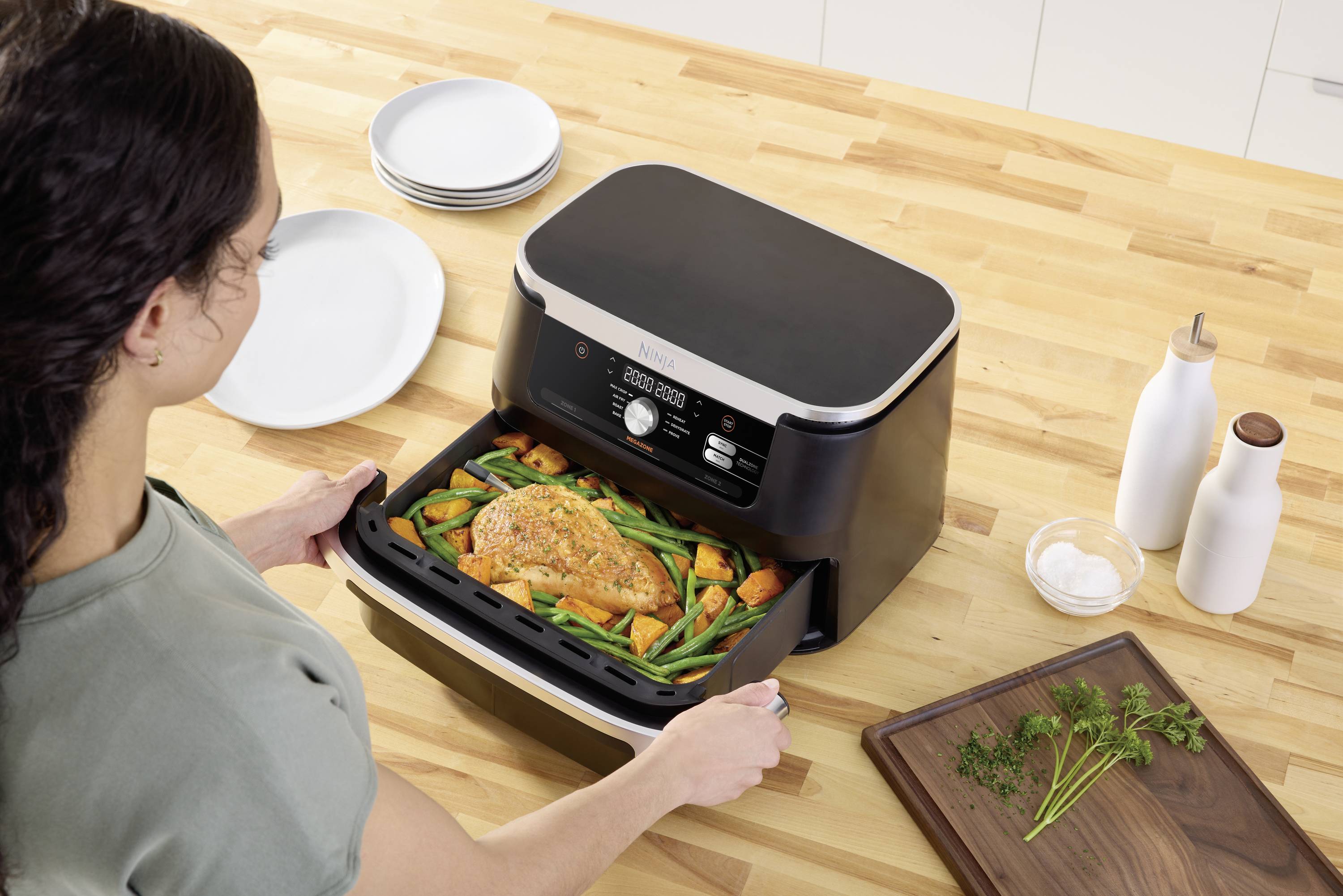 Eine Frau öffnet eine Heißluftfritteuse mit einem gebratenen Hühnchen und Gemüse auf einem Holztisch. Teller und Gewürze sind nebenan.
