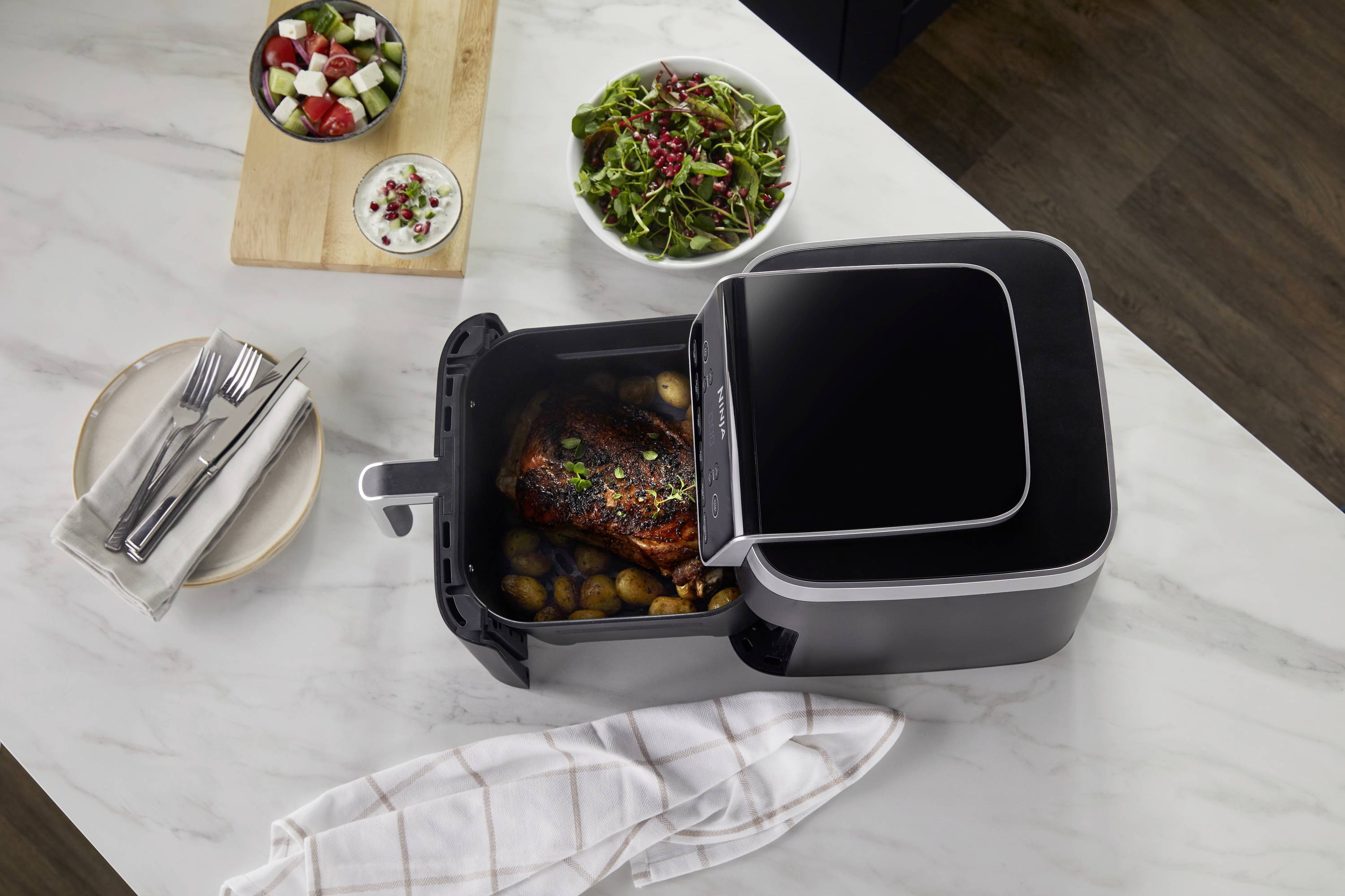 Eine Heißluftfritteuse mit geöffnetem Korb zeigt gebratenes Fleisch und Kartoffeln. Nebenbei steht ein frischer Salat und Besteck.