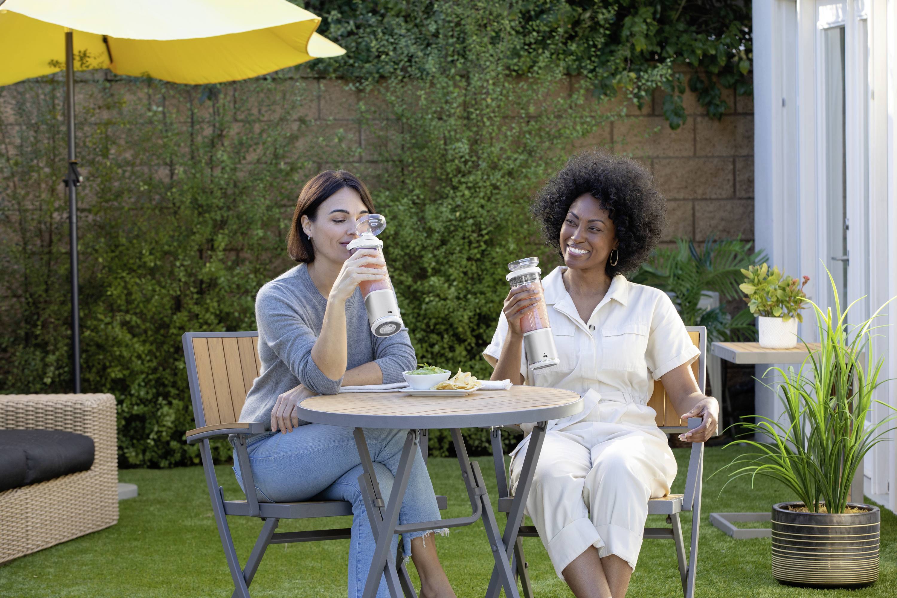 Zwei Frauen sitzen im Garten an einem Tisch und trinken aus Trinkflaschen. Es ist sonnig, und im Hintergrund sind Pflanzen zu sehen.