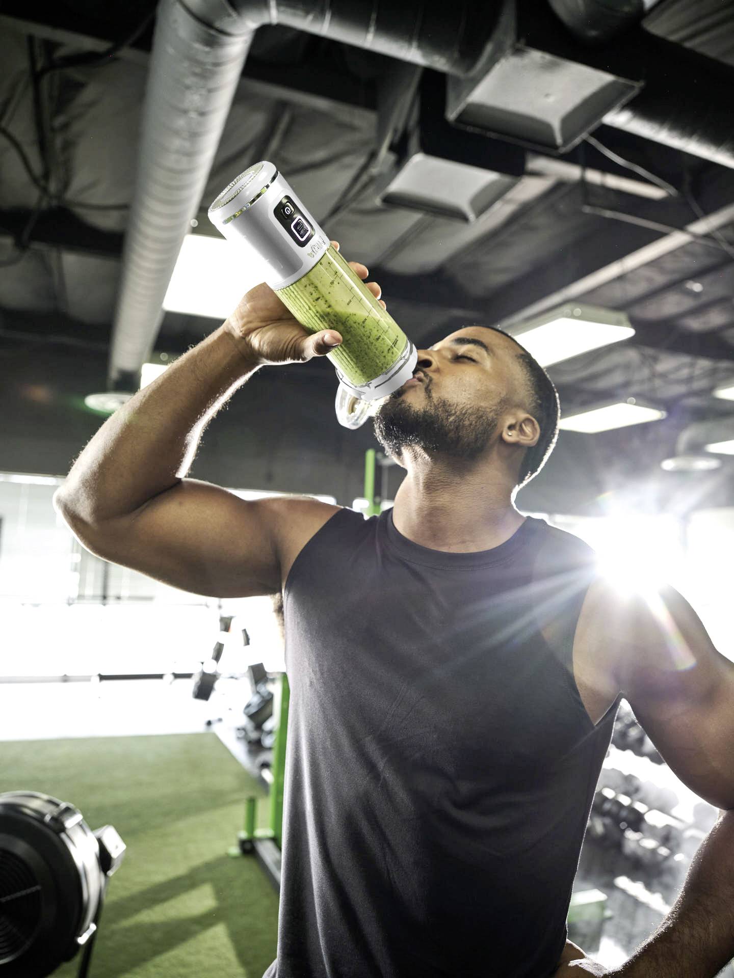 Ein Mann im Fitnessstudio trinkt aus einer Flasche mit einem grünen Getränk. Sonnenlicht strahlt von hinten. Fitnessgeräte sind sichtbar.
