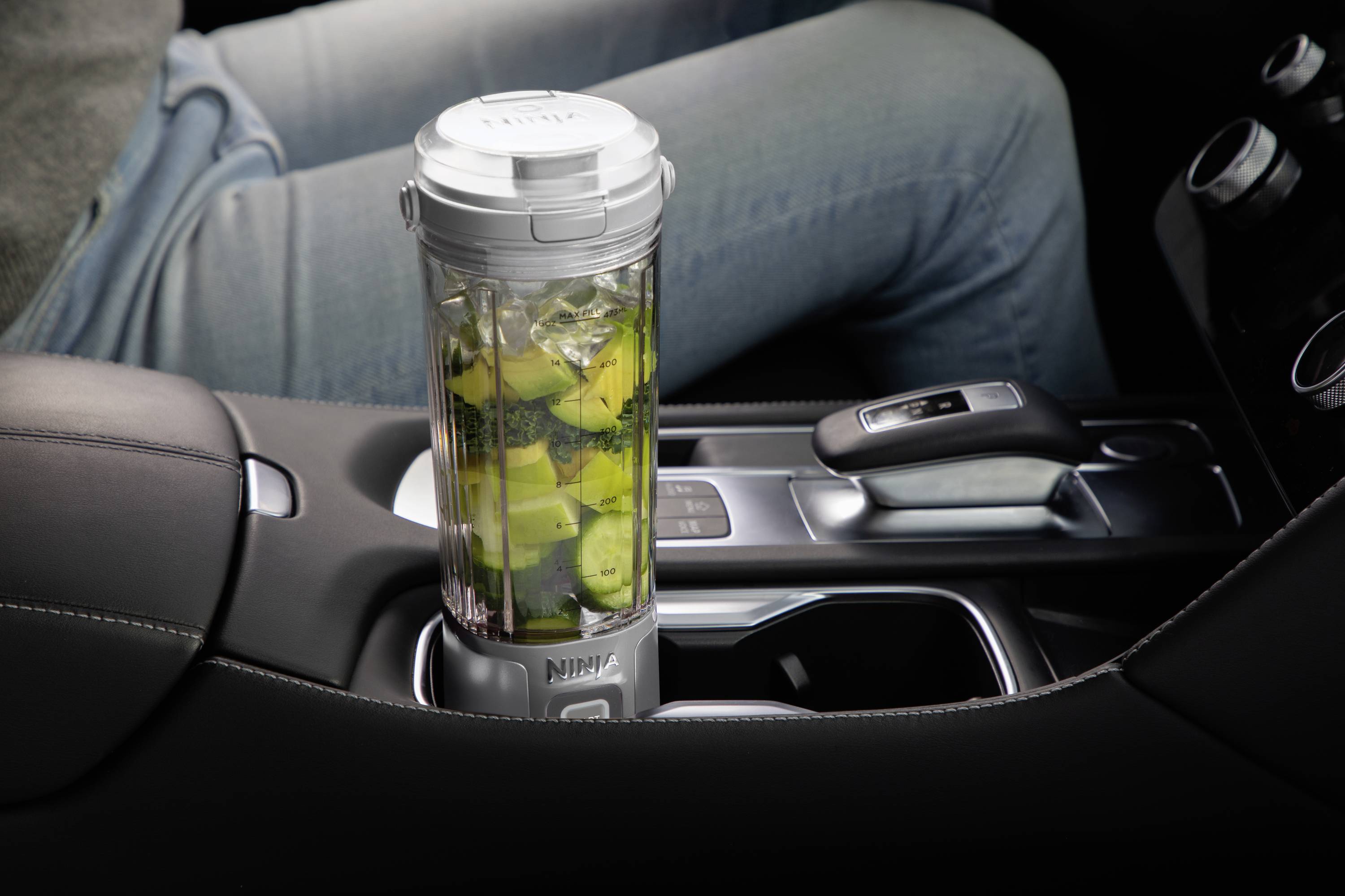 Ein tragbarer Mixer gefüllt mit Obst und Gemüse steht in der Mittelkonsole eines Autos. Eine Person sitzt am Steuer.