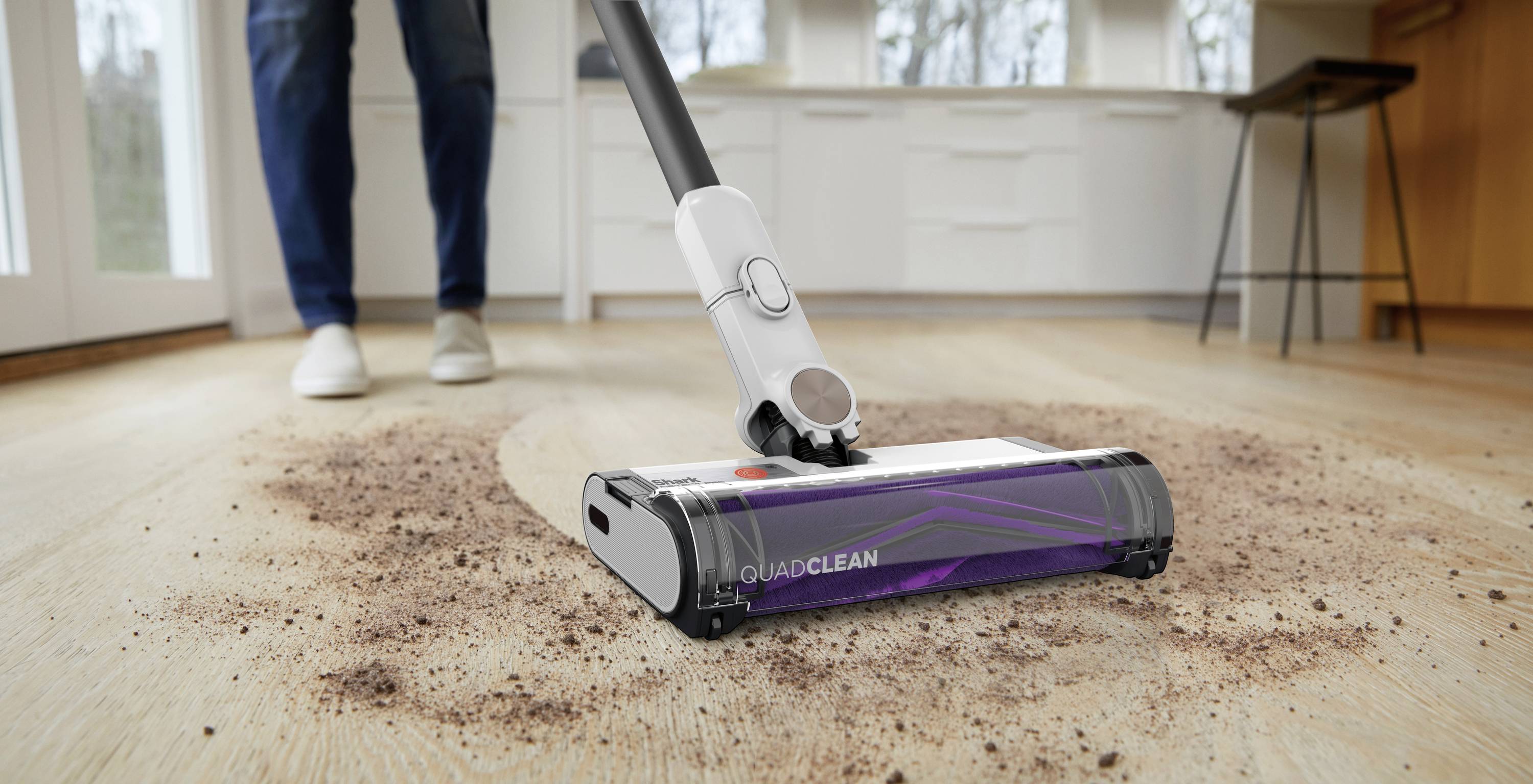 Person saugt mit einem lila "Quadclean"-Staubsauger Schmutz vom Holzboden in einer hellen Küche auf.