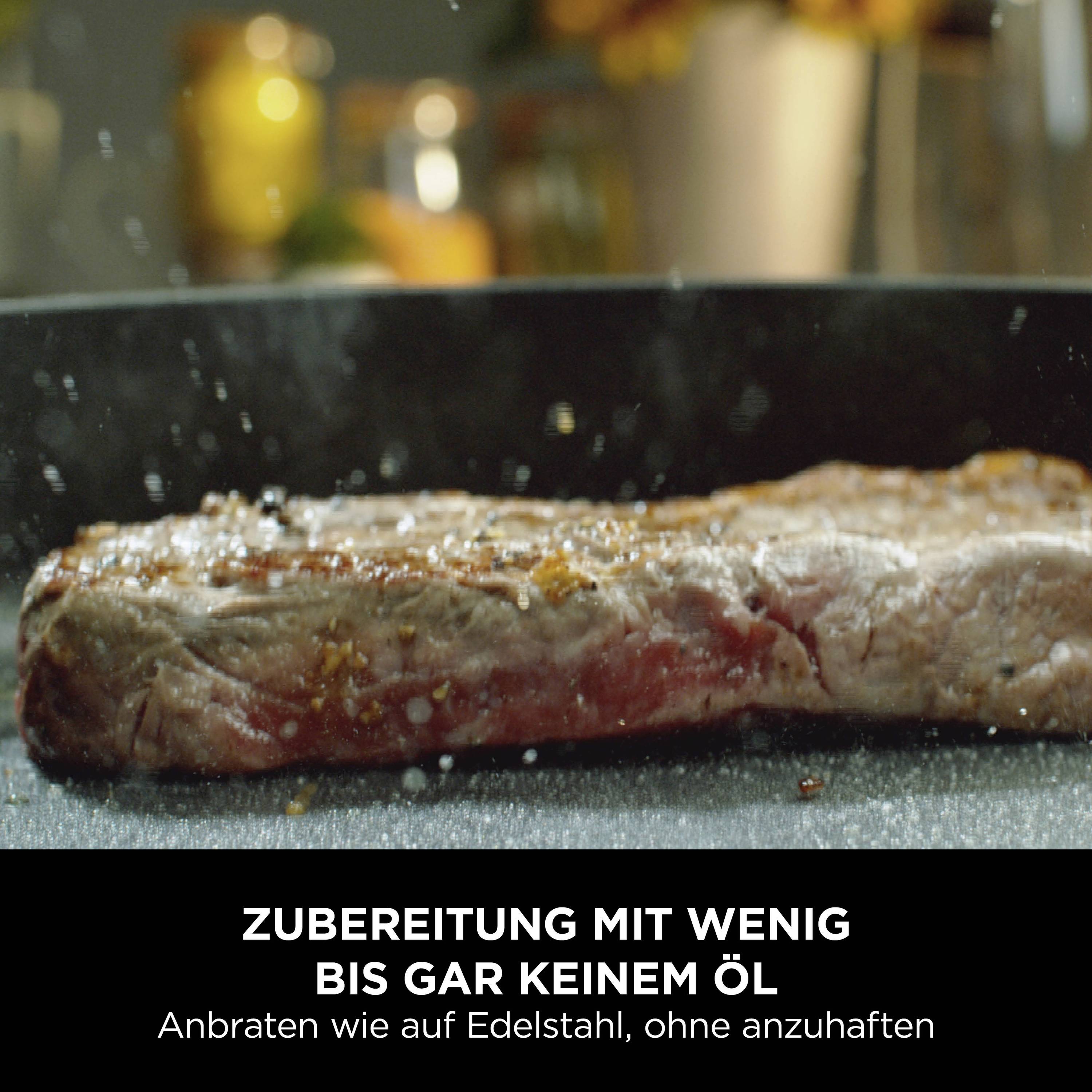 Ein Stück Fleisch brät in einer schwarzen Pfanne. Darunter steht: 'Zubereitung mit wenig bis gar keinem Öl, anbraten wie auf Edelstahl, ohne anzuhäften'.
