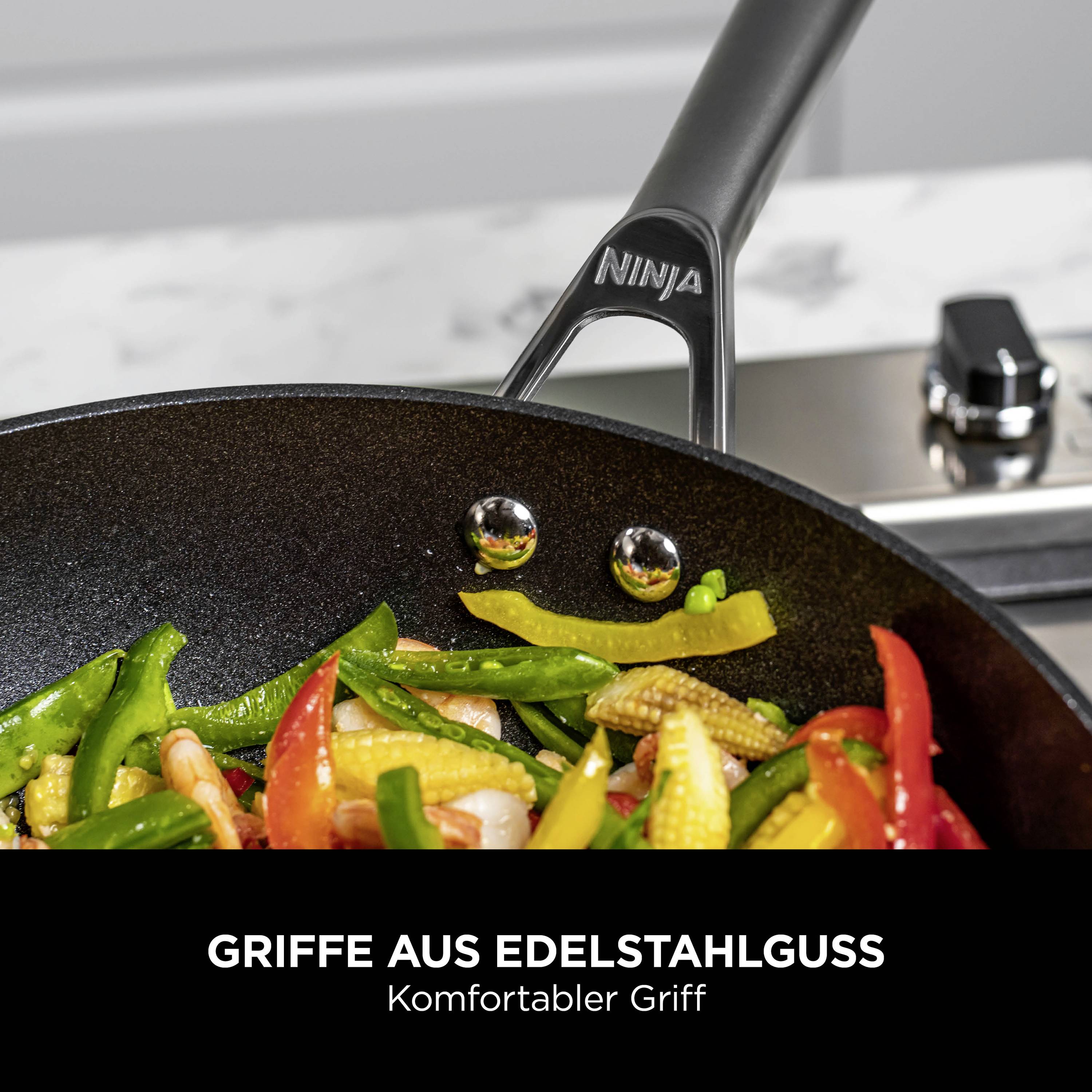 Pfanne auf Herd mit buntem Gemüse. Aufschrift 'GRIFFE AUS EDELSTAHLGUSS, Komfortabler Griff'. Markierung 'NINJA'.