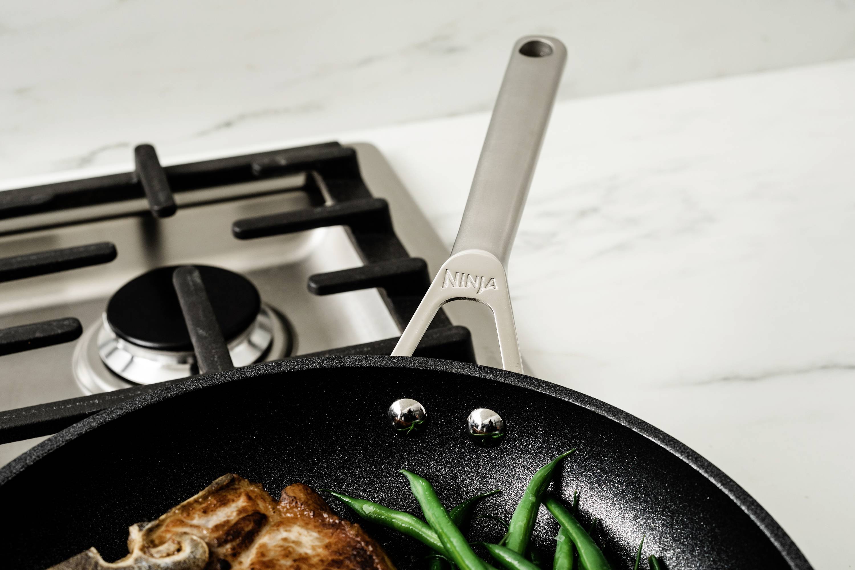 Eine Bratpfanne mit grünem Gemüse und einem Stück Fleisch auf einem Herd neben einem Gasbrenner.