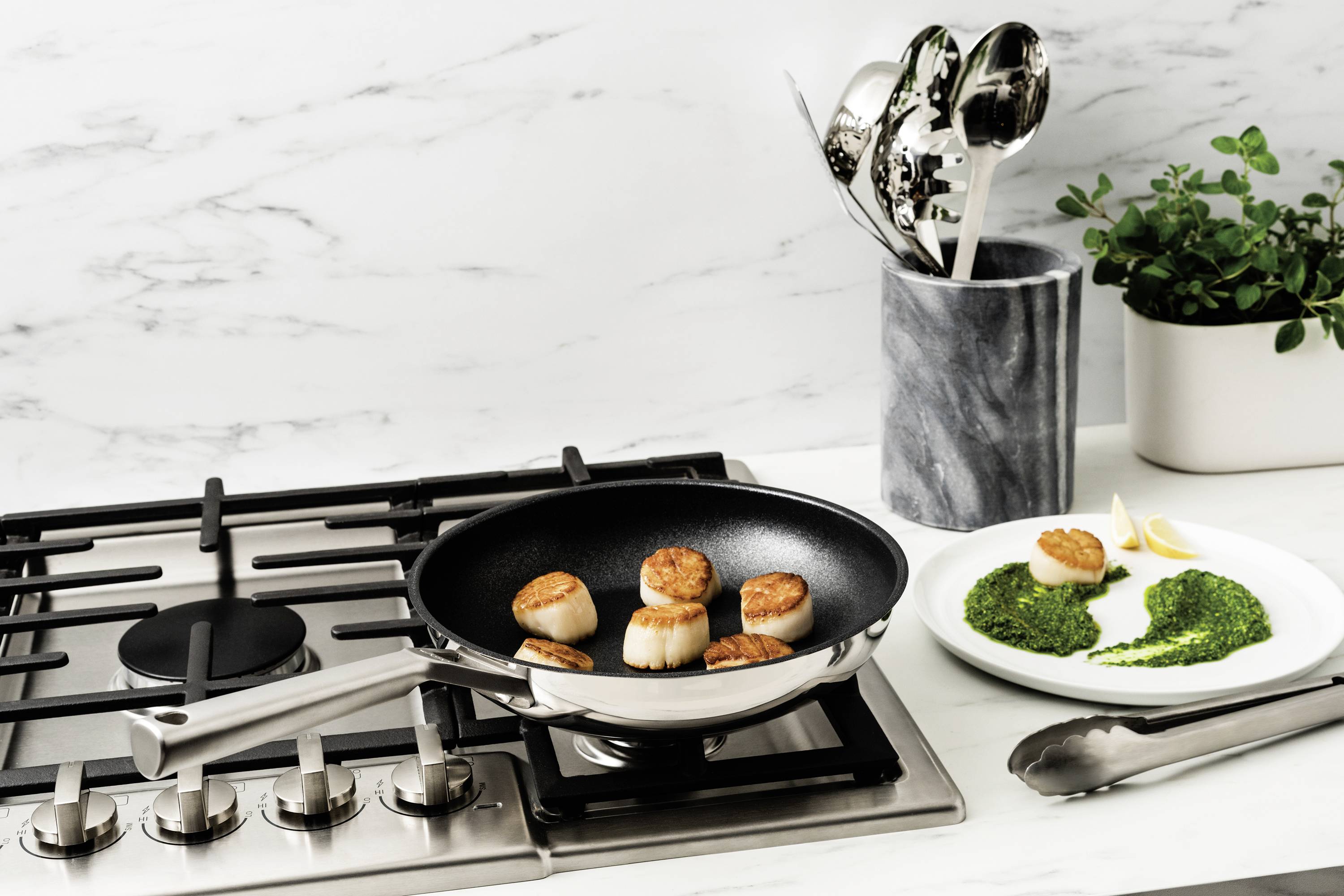 Eine Pfanne mit gebratenen Jakobsmuscheln auf einem Gasherd, daneben ein Teller mit Pesto und Zitronenscheiben. Dekoratives Küchenset.