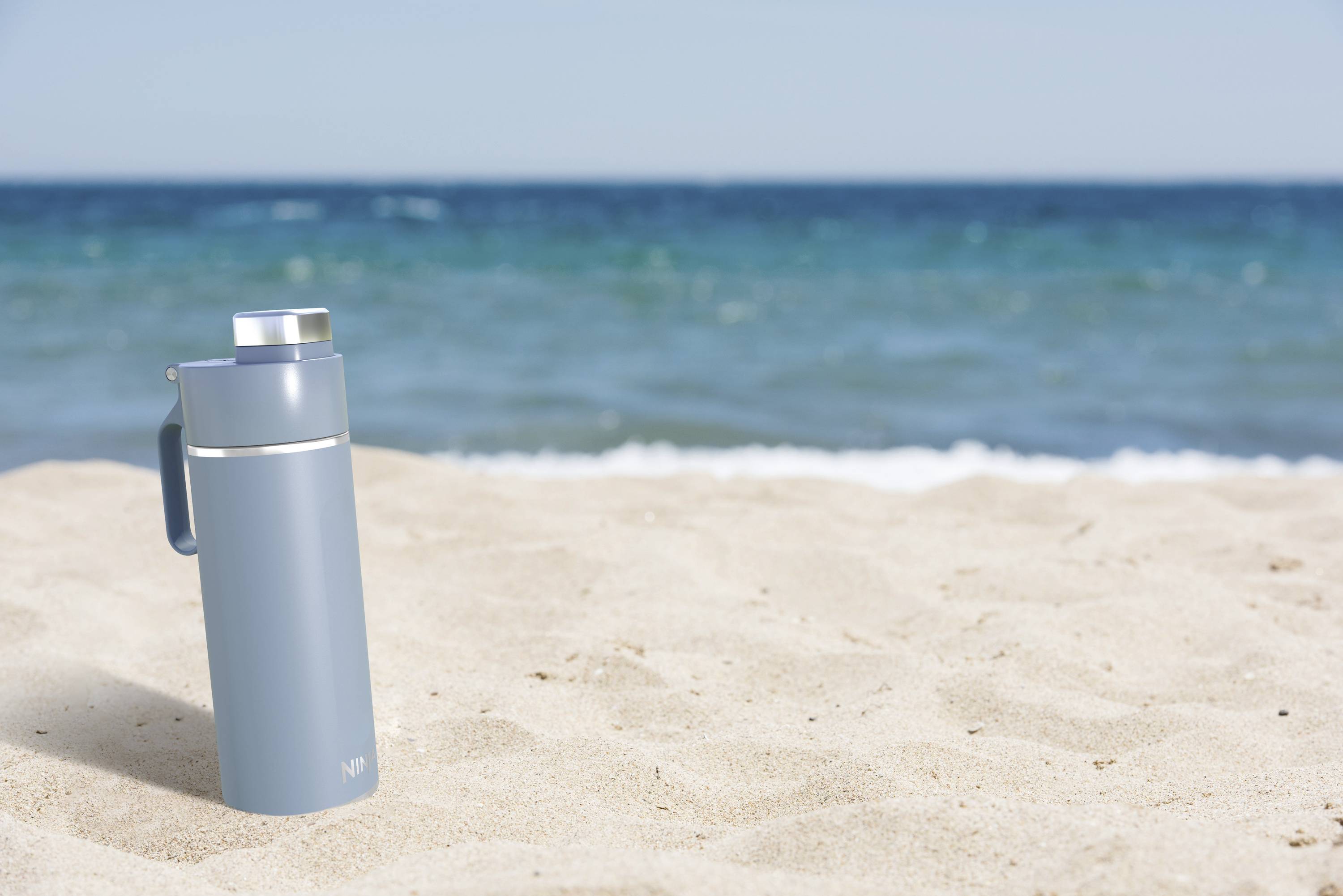 Eine blaue Thermosflasche steht im Sand am Strand. Im Hintergrund sind Wellen des Meeres und der klare Himmel zu sehen.