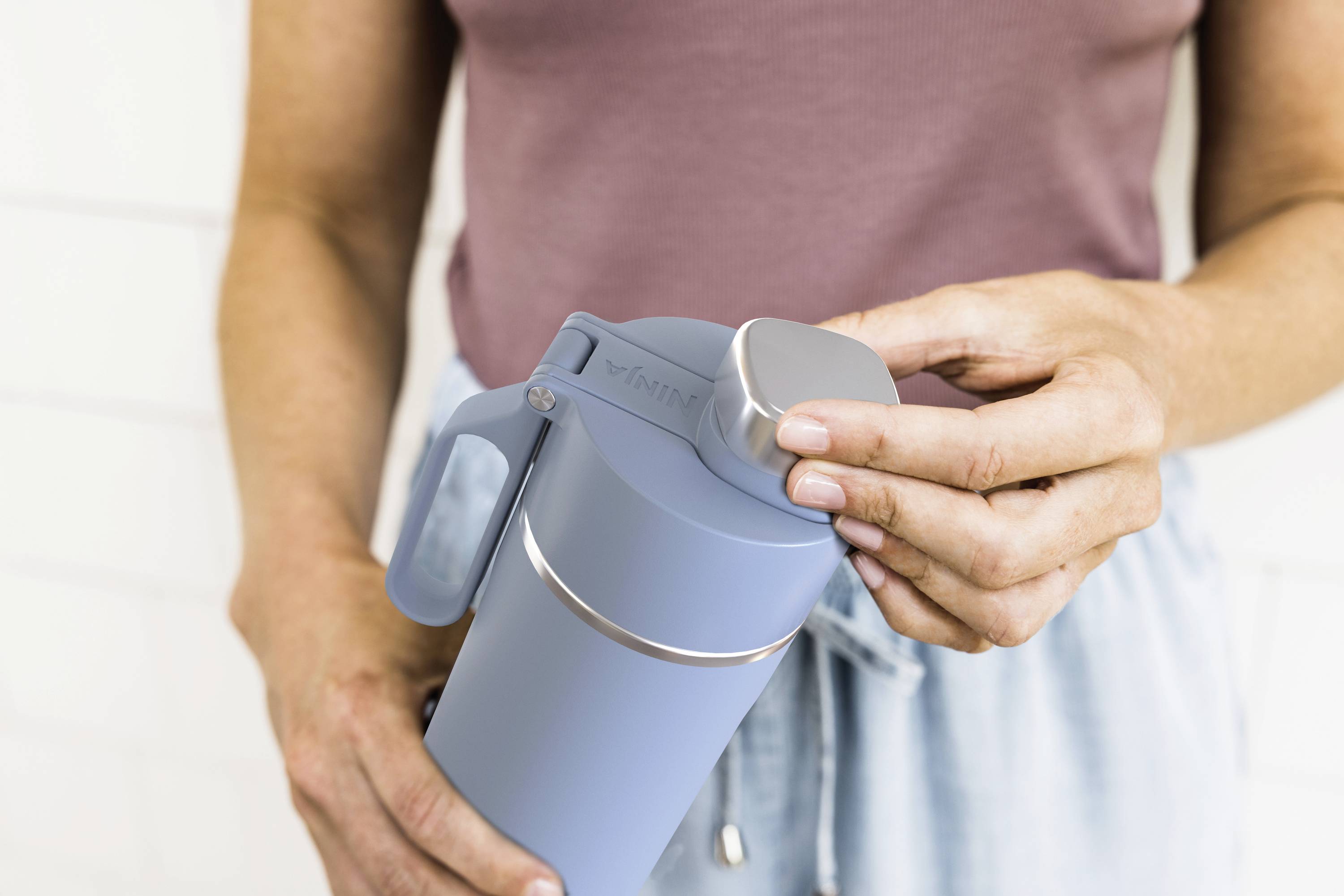 Eine Person hält eine hellblaue Wasserflasche und öffnet den Deckel. Sie trägt ein ärmelloses, rosa Oberteil und eine blaue Hose.