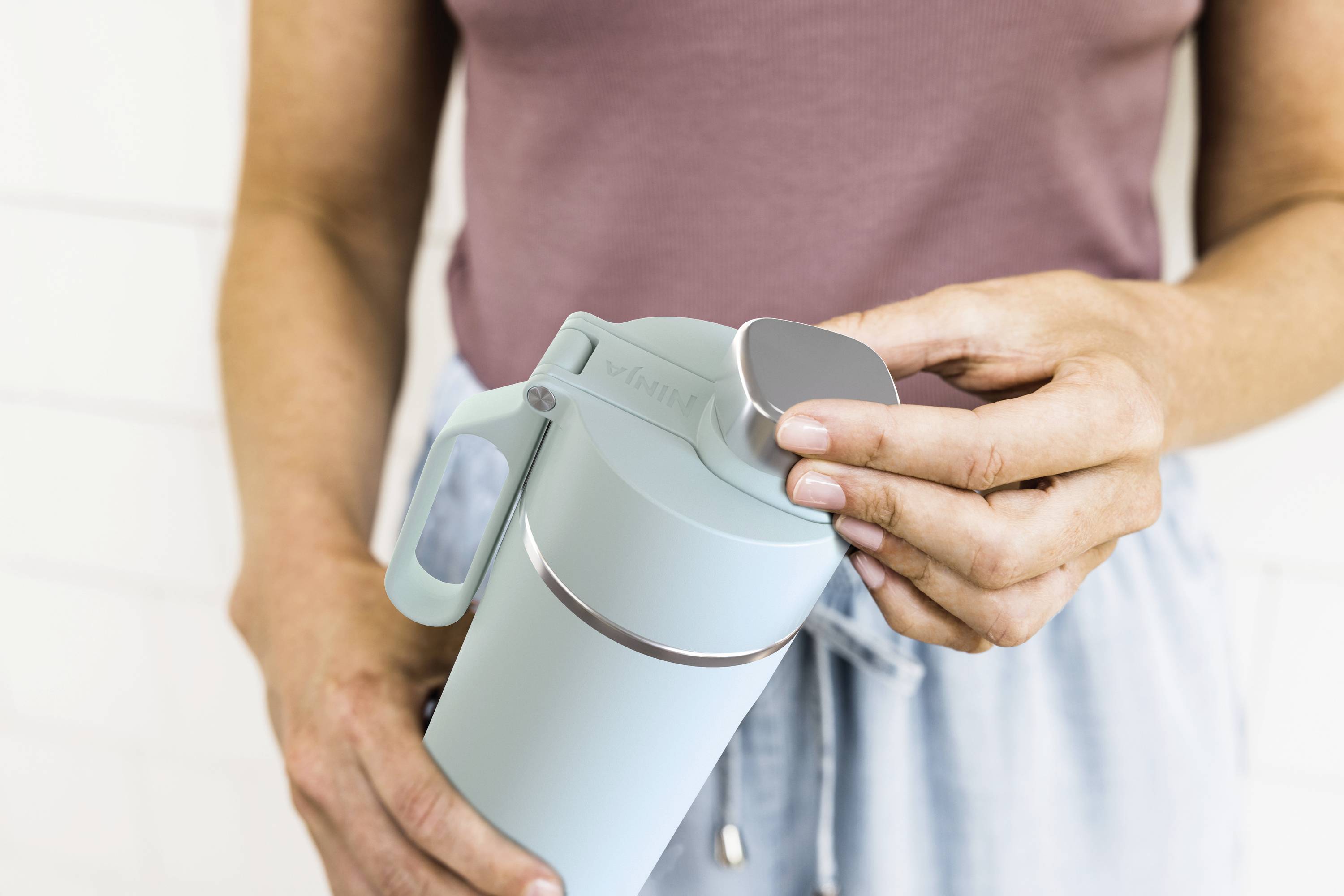 Eine Person hält eine hellblaue Thermosflasche, öffnet den Deckel und zeigt eine Funktion der Flasche in einer entspannten Umgebung.