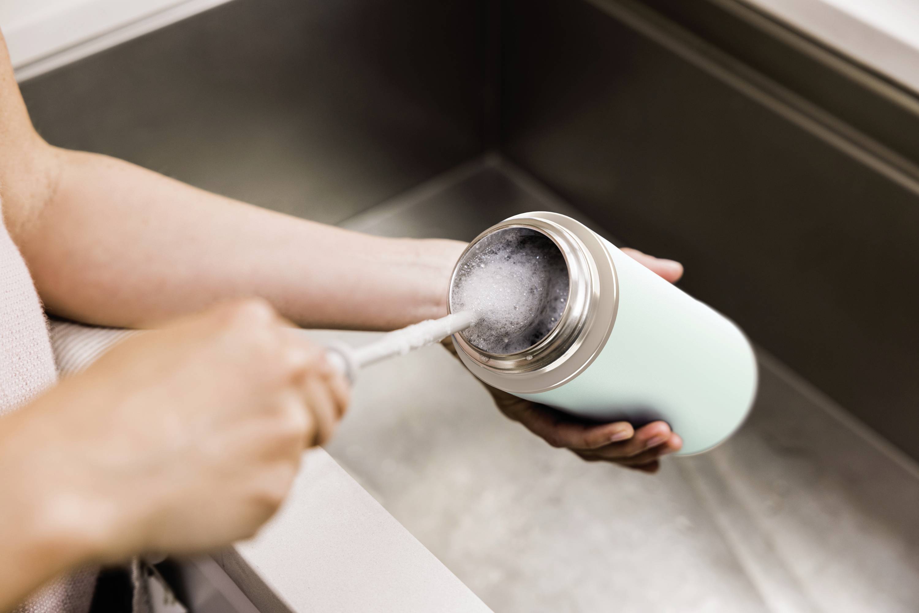 Eine Person reinigt eine hellblaue Thermoskanne im Waschbecken mit einer Reinigungsbürste und Wasser.