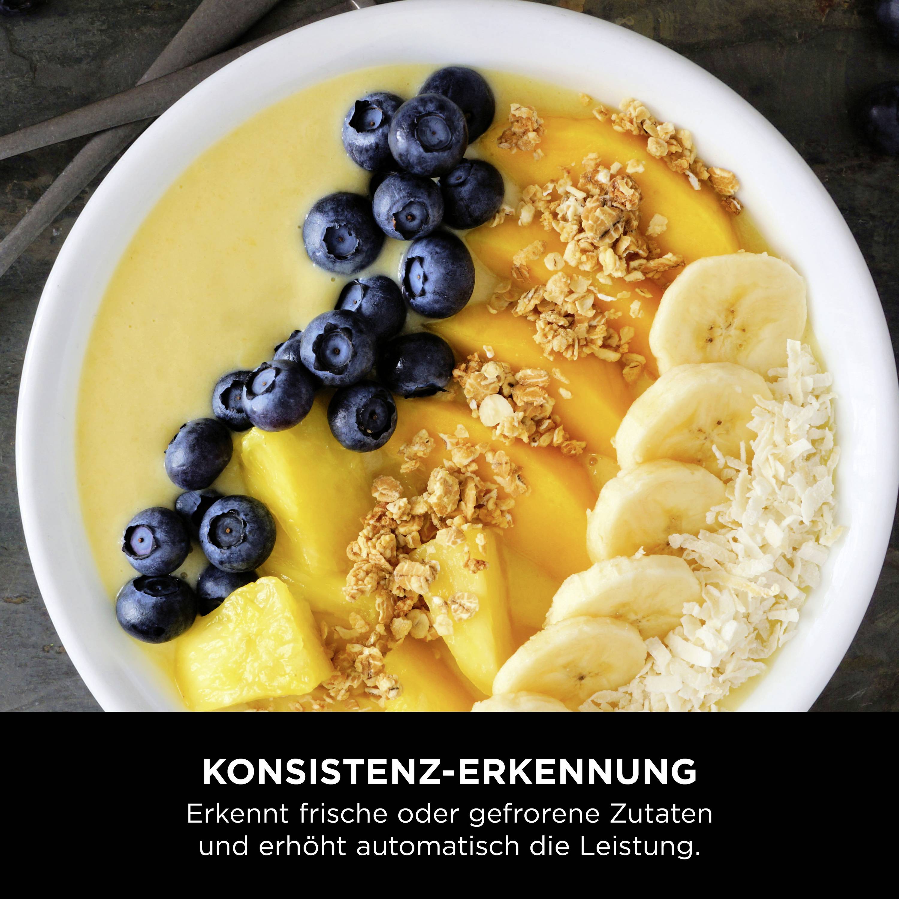 Eine Schale mit gelber Smoothie-Basis, bedeckt mit Blaubeeren, Mangostücken, Bananenscheiben und Müsli. Darunter Text: 'Konsistenz-Erkennung: Erkennt frische oder gefrorene Zutaten und erhöht automatisch die Leistung'.