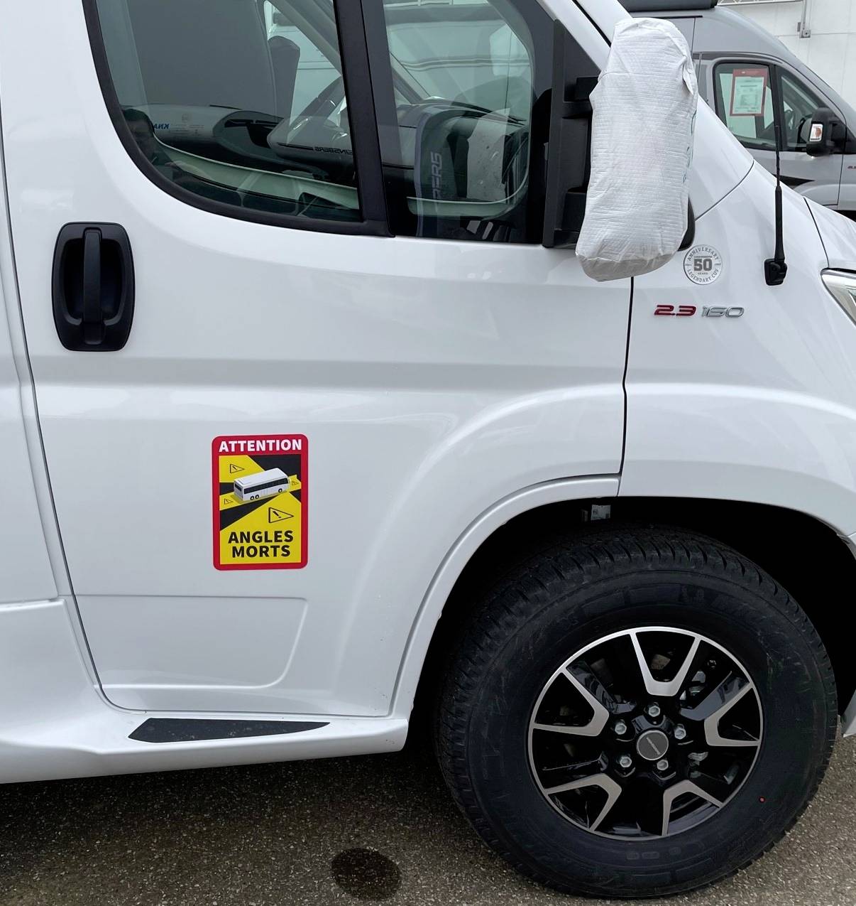 Ein weißer Transporter mit einem Aufkleber 'Attention Angles Morts', der vor toten Winkeln warnt. Der Seitenspiegel ist geschützt.