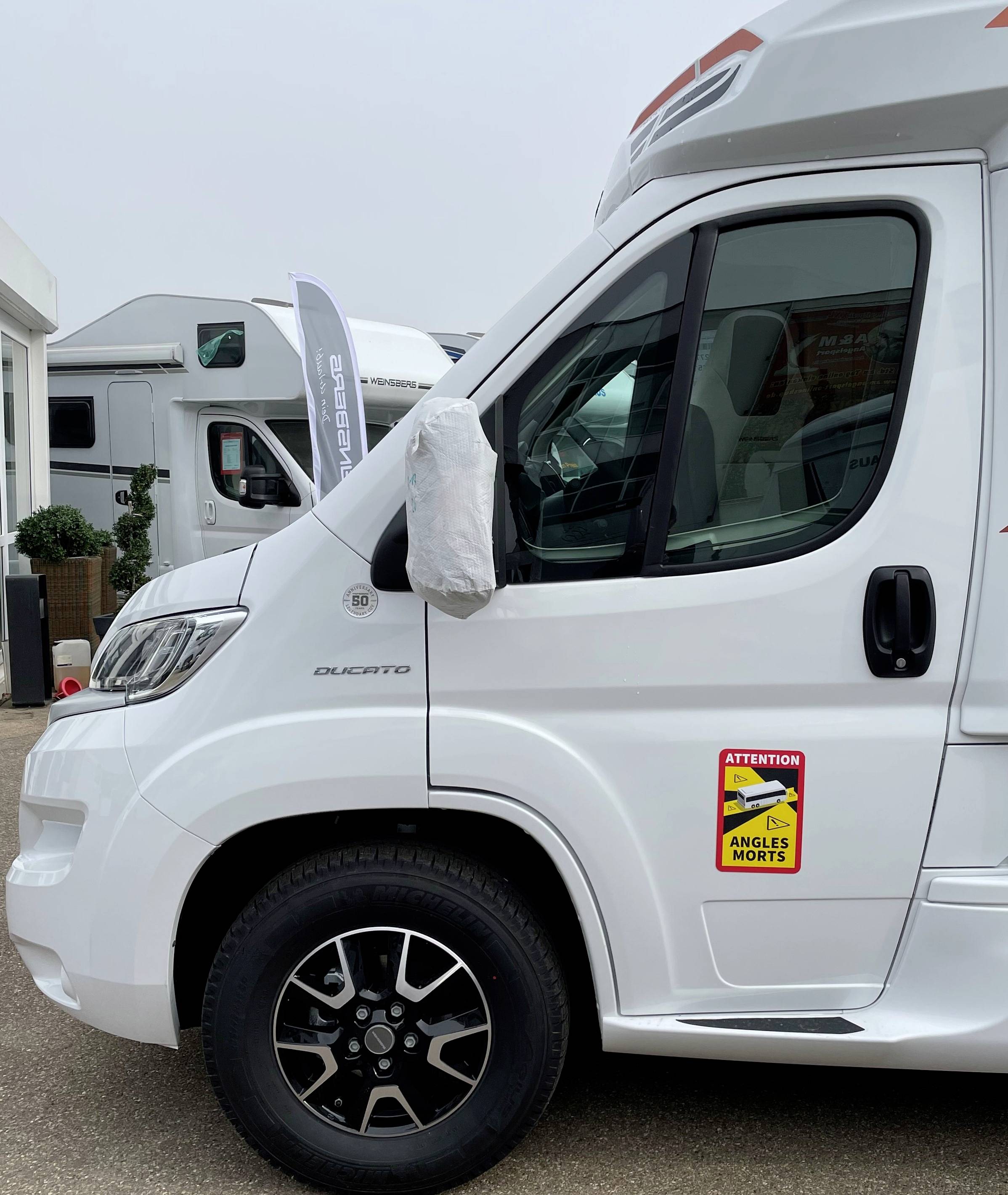 Ein weißes Wohnmobil mit einem Aufkleber, auf dem steht 'Attention - Angles Morts', parkt in einer Wohnmobil-Ausstellung.
