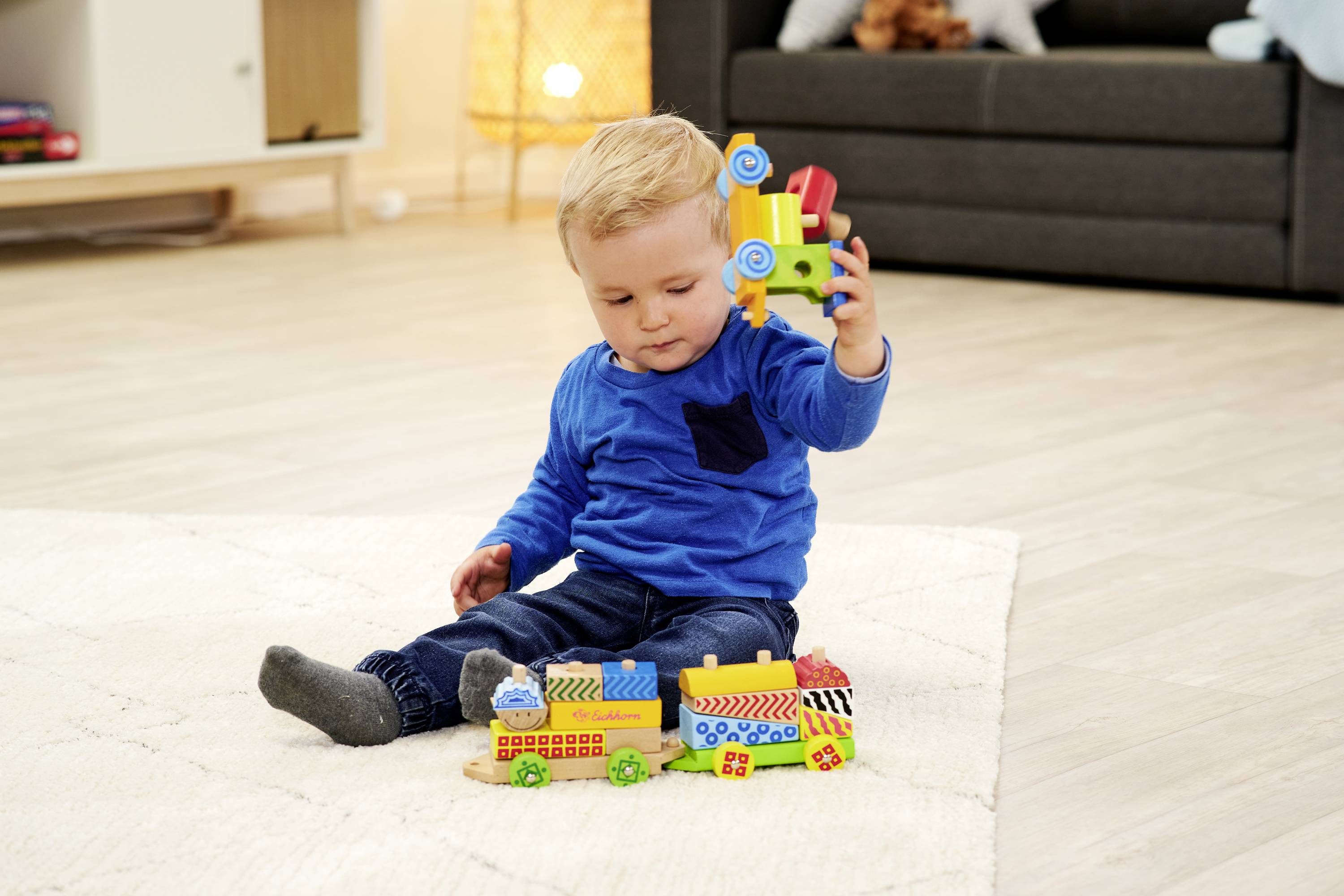 Ein Kleinkind sitzt auf dem Boden und spielt mit bunten Bauklötzen in einem Wohnzimmer. Der Hintergrund zeigt ein Sofa und eine Lampe.