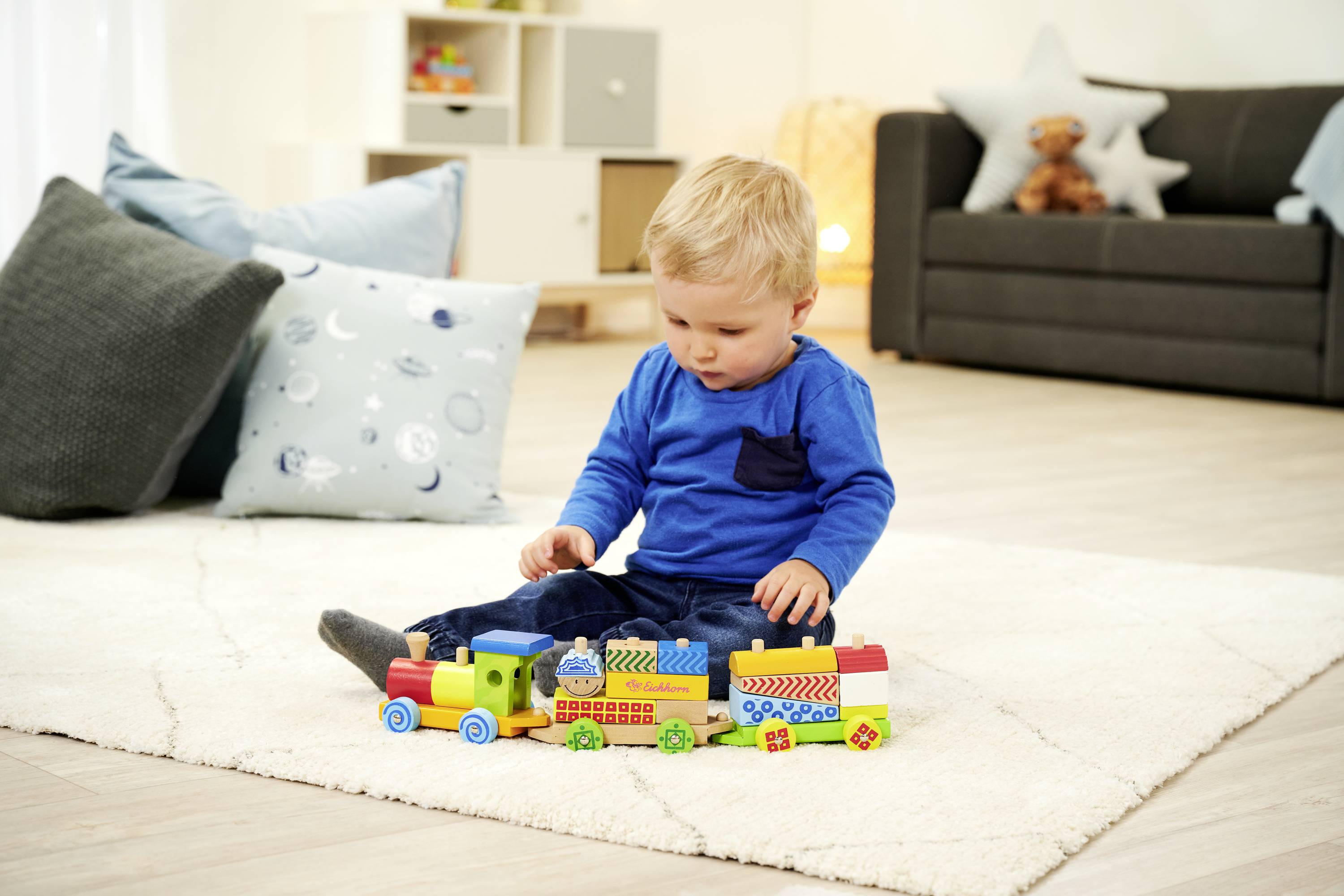 Kleinkind sitzt auf Teppich und spielt mit buntem Spielzeugzug; im Hintergrund ein Sofa, Kissen und Regale mit Spielzeug.