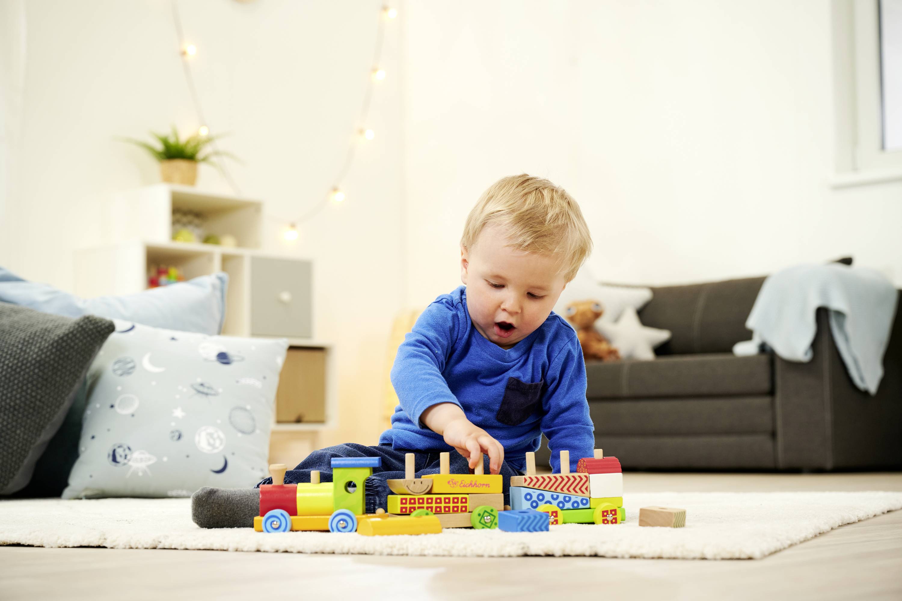Ein Kleinkind spielt auf einem weißen Teppich mit buntem Holzspielzeugzug in einem Wohnzimmer mit grauem Sofa im Hintergrund.