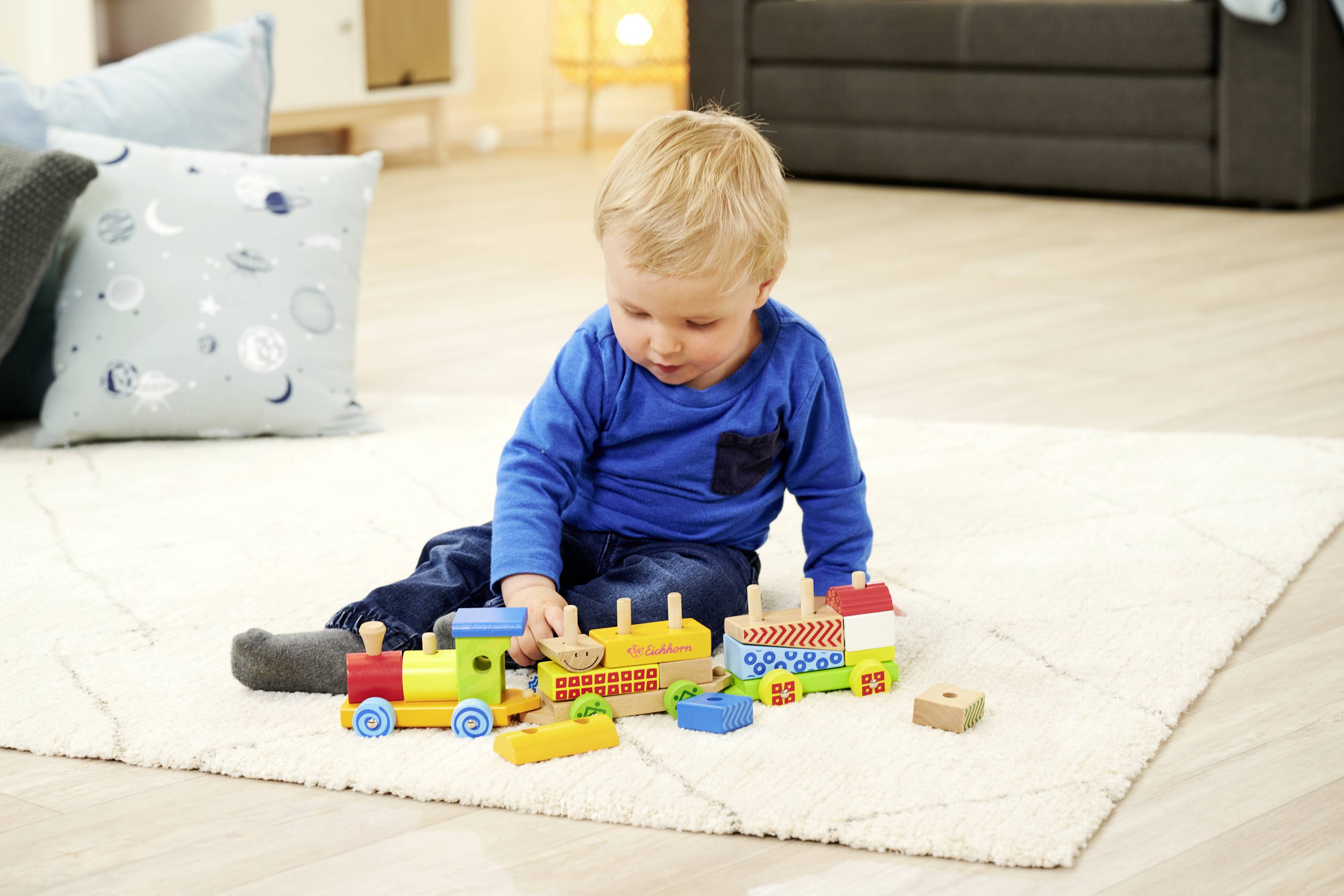 Ein Kleinkind sitzt auf einem Teppich und spielt mit bunten Holzbausteinen, die zu einer Zugform zusammengebaut sind. Im Hintergrund sind ein Sofa und Kissen zu sehen.