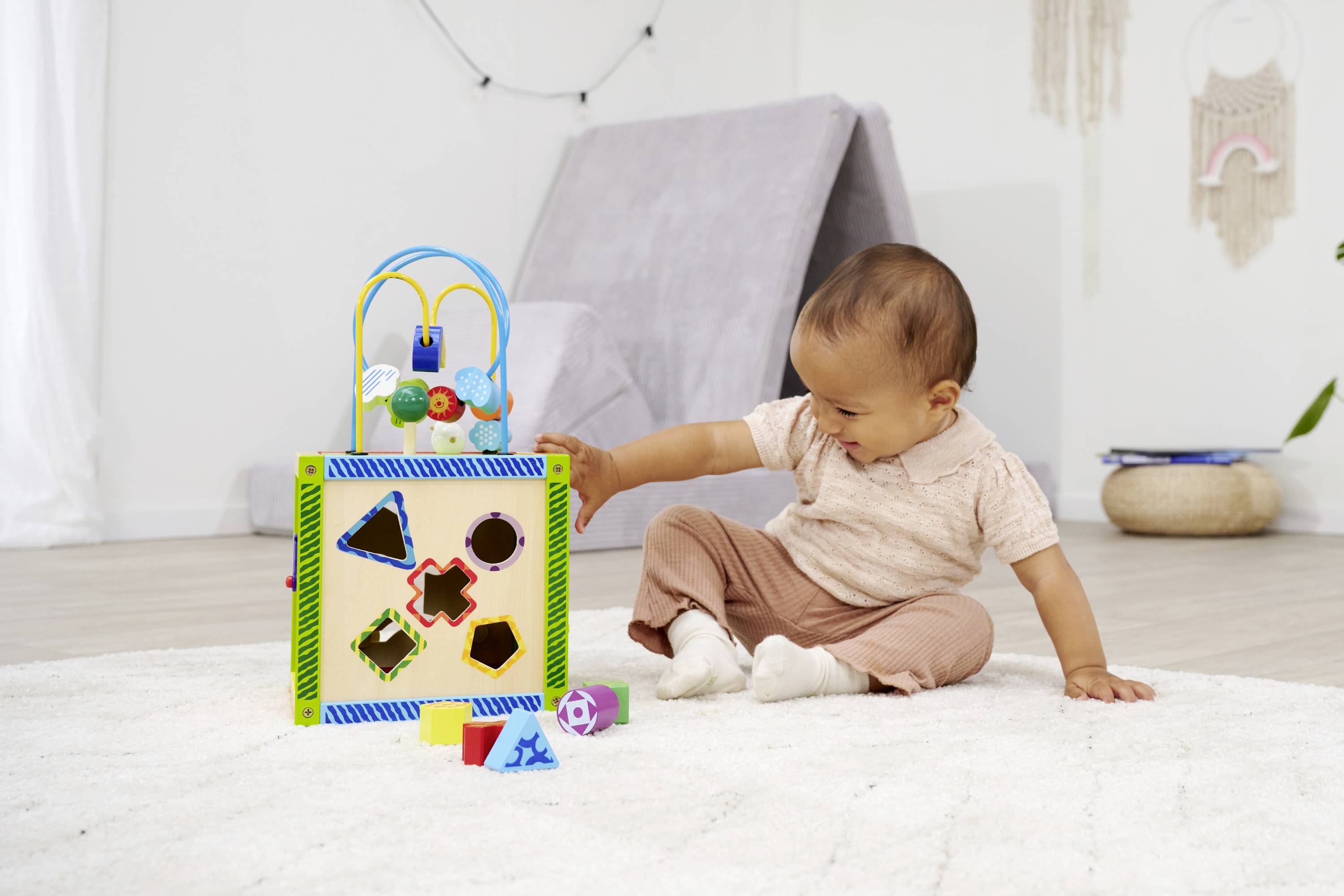 Ein Baby sitzt auf einem Teppich und spielt mit einem bunten Spielzeug, das Formen hat. Der Raum ist hell und gemütlich eingerichtet.