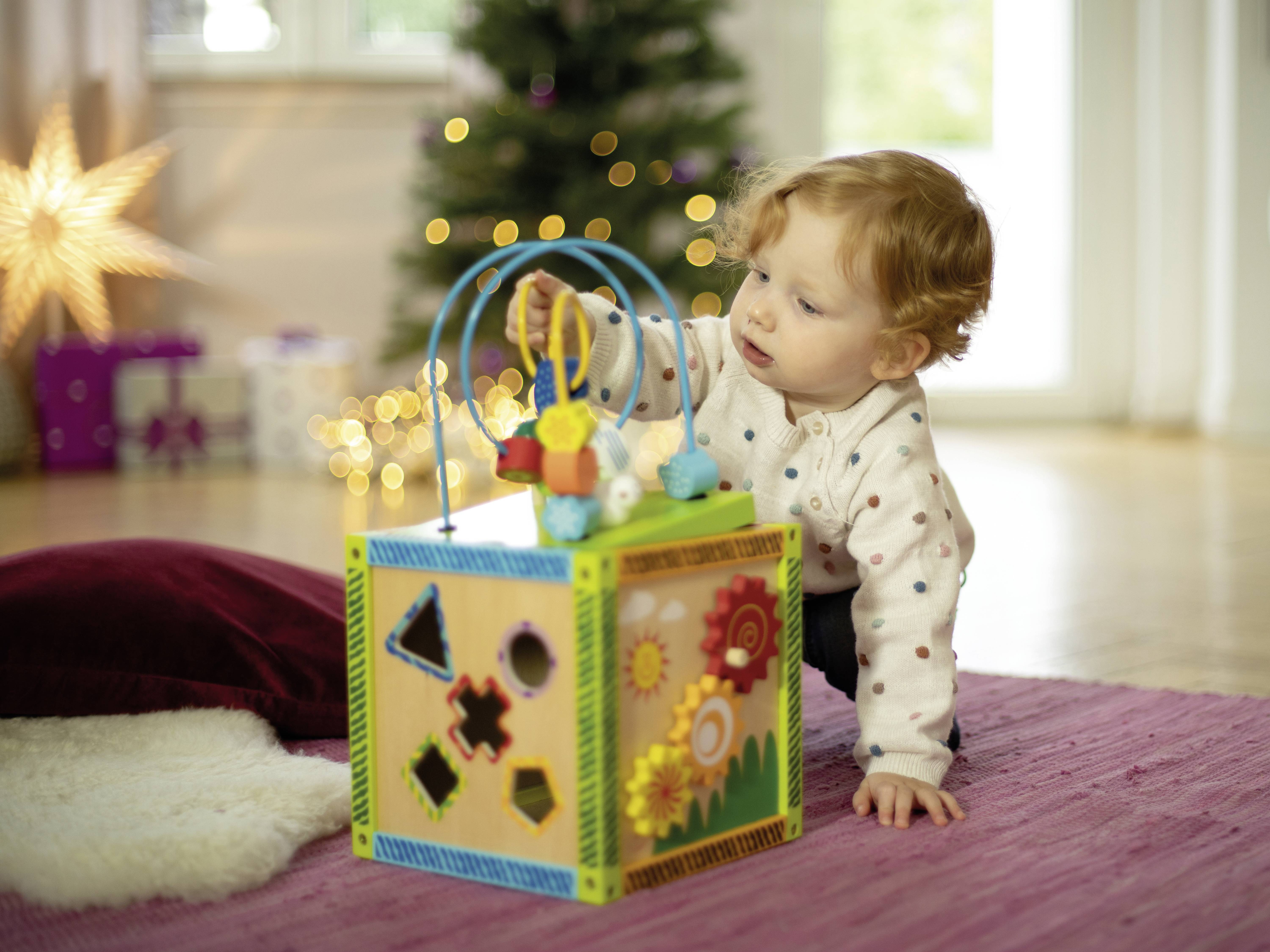 Ein Kleinkind spielt auf einem Teppich mit einem Spielwürfel, der Formen zum Einsetzen und bunte Blumenmuster hat. Im Hintergrund steht ein geschmückter Weihnachtsbaum.