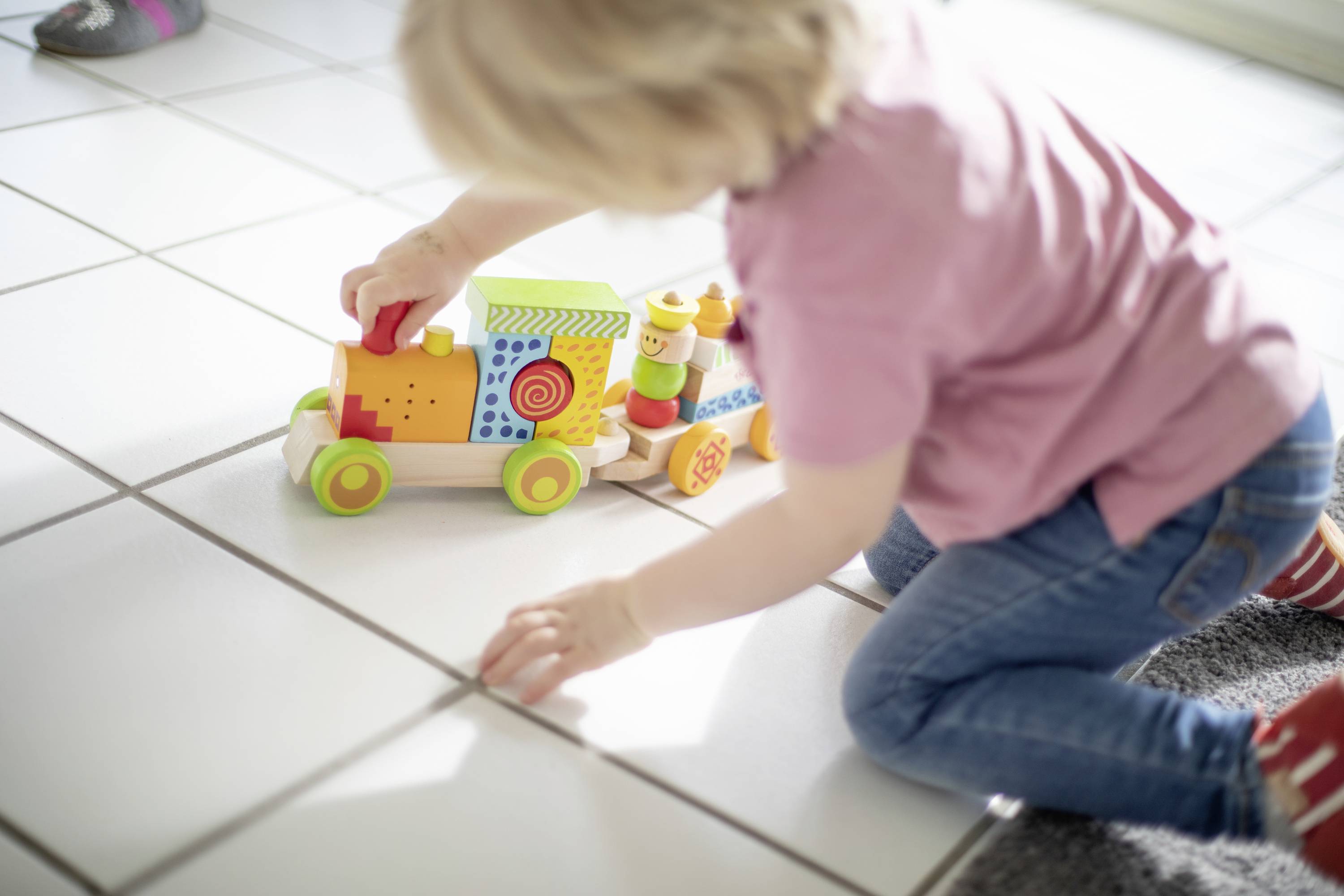 Ein kleines Kind spielt auf dem Boden mit einem bunten Holzspielzeugzug. Es trägt ein rosa T-Shirt und blaue Jeans.