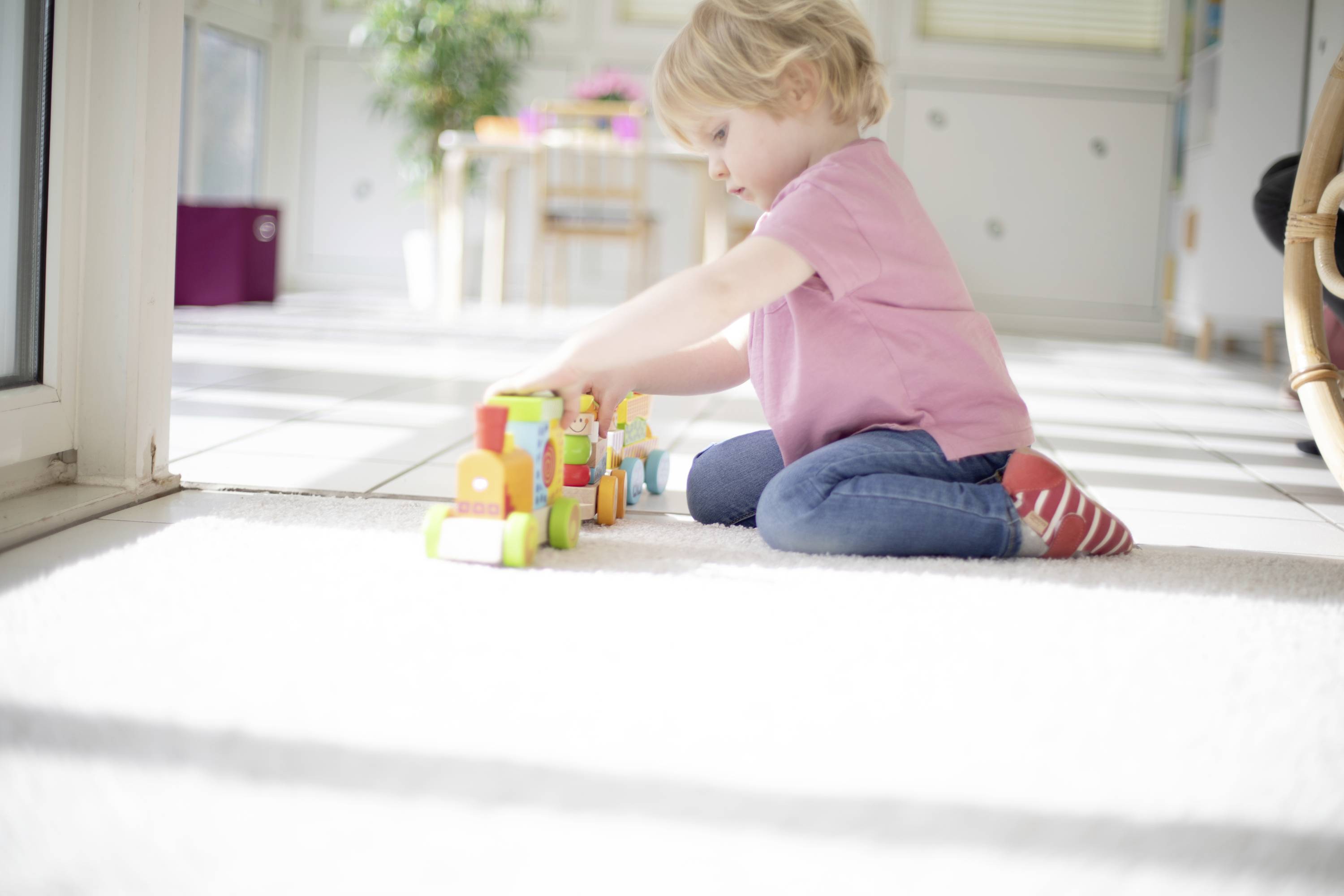 Ein kleines Kind sitzt auf dem Boden und spielt konzentriert mit bunten Holzbausteinen in einem hellen Raum.