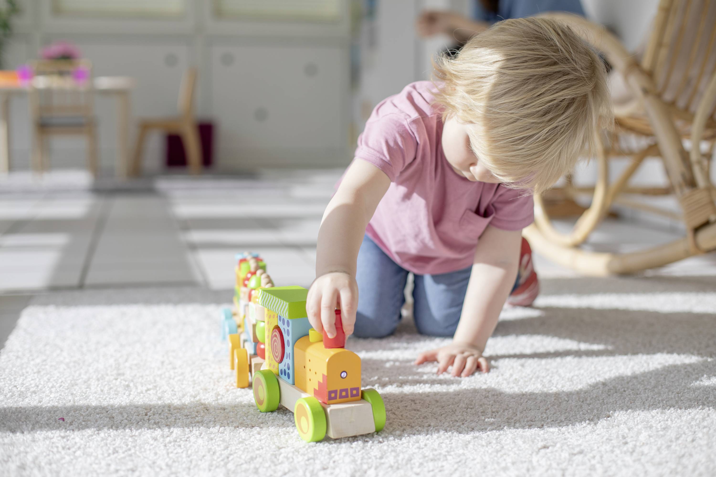 Kleinkind spielt auf dem Teppich mit einem bunten Holzspielzeugzug. Im Hintergrund sind Möbel und ein Schaukelstuhl sichtbar.