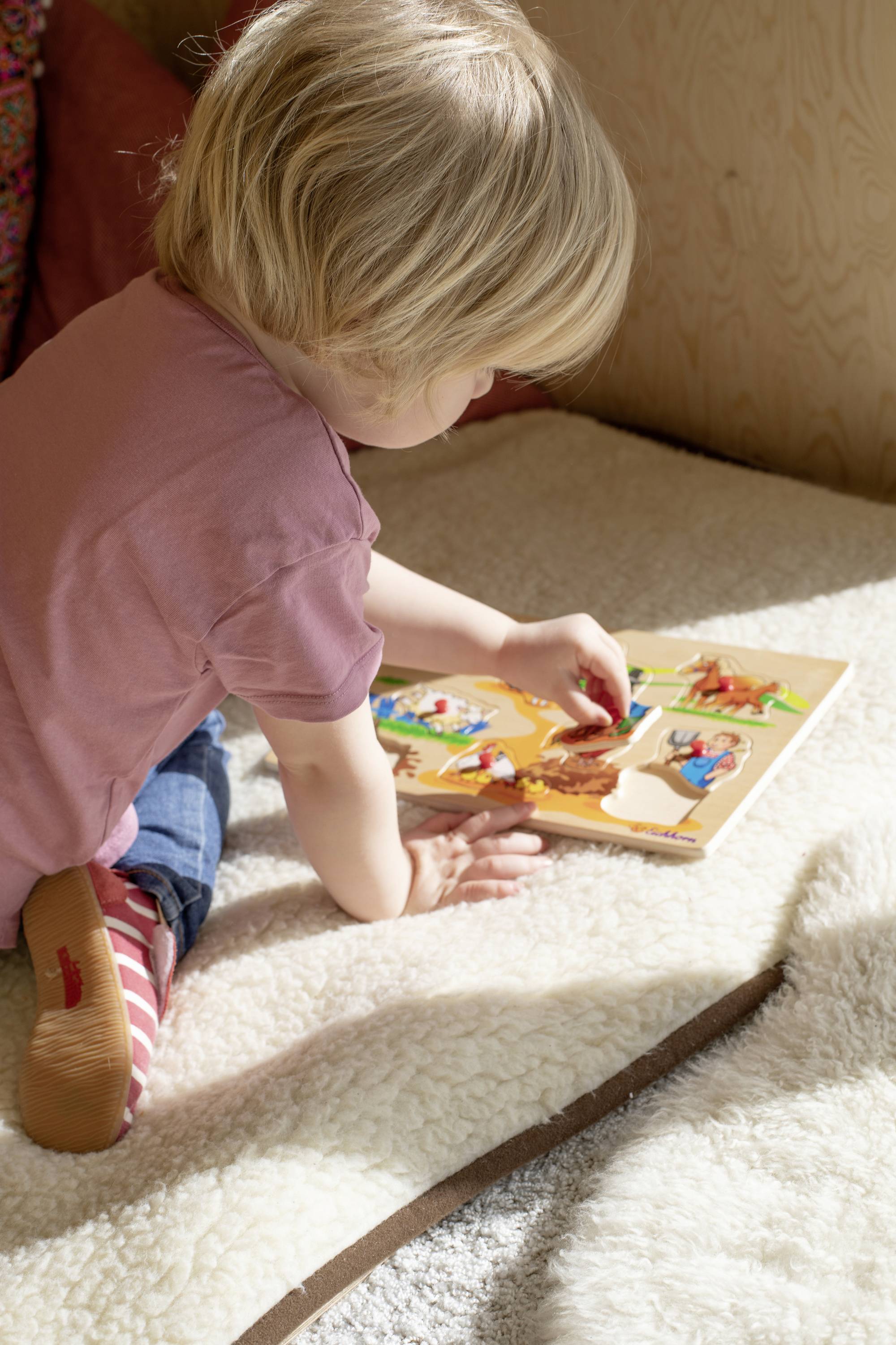 Ein kleines Kind spielt auf dem Boden mit einem Holzpuzzle. Das Kind sitzt auf einer weichen, hellen Decke in einem gut beleuchteten Raum.