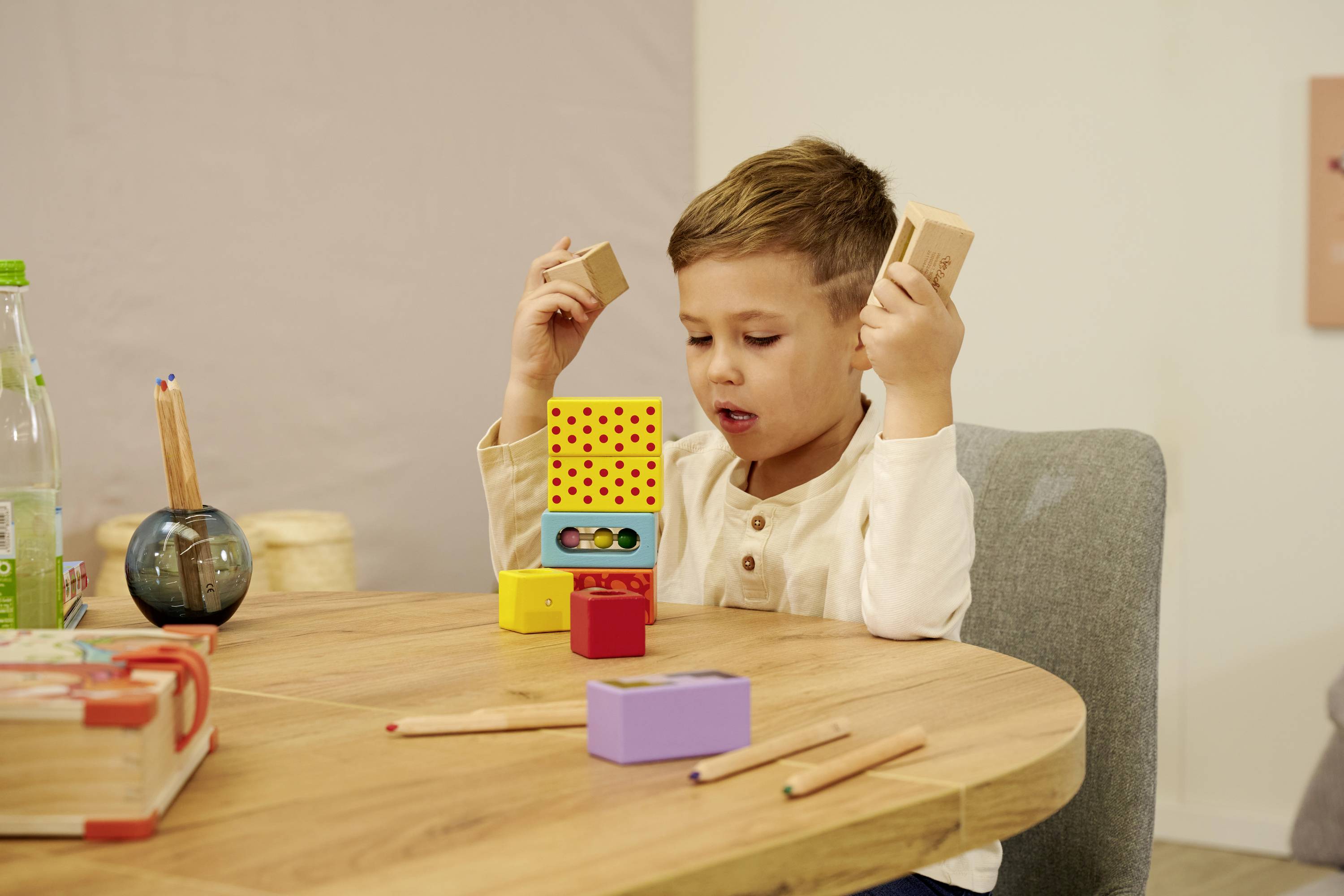 Ein Kind sitzt an einem Holztisch und spielt mit bunten Holzklötzen. Im Hintergrund sind Buntstifte und eine Wasserflasche zu sehen.