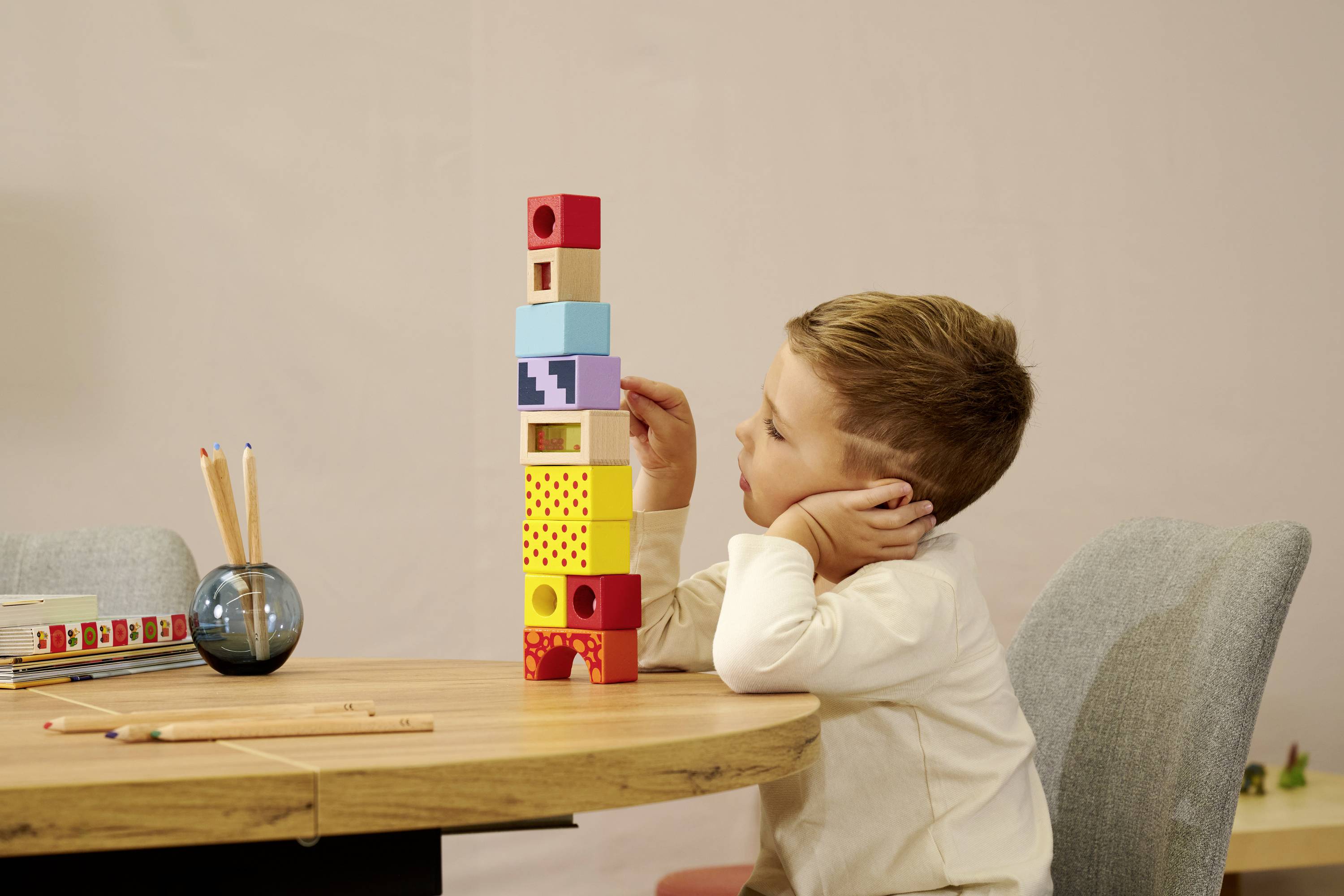 Ein Kind sitzt an einem Tisch und baut einen hohen Turm aus bunten Holzklötzen. Neben ihm stehen Buntstifte in einem Glas.