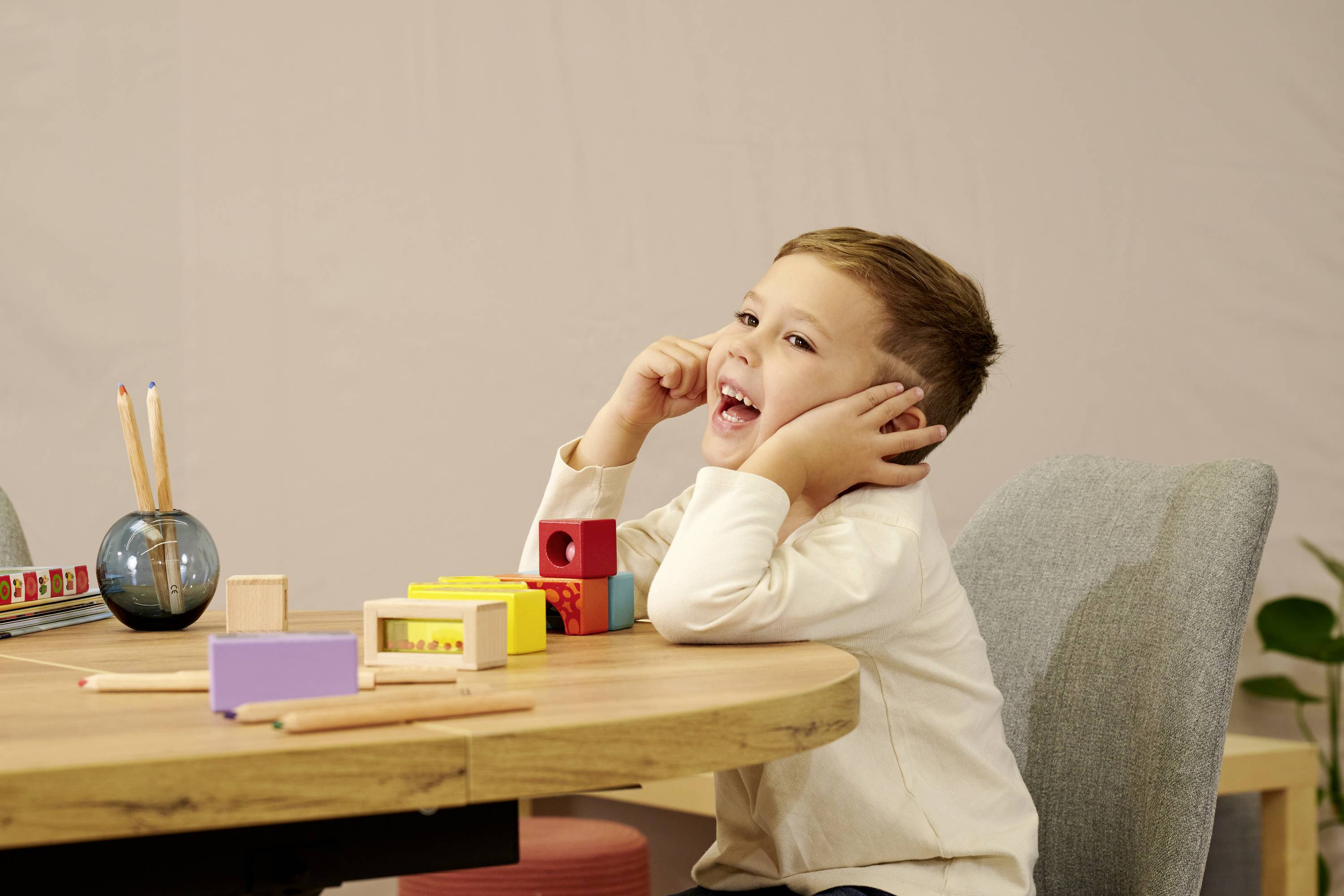 Ein lachender Junge sitzt an einem Tisch mit Spielklötzen und Stiften. Er trägt ein weißes Hemd und seine Hände berühren seine Ohren.