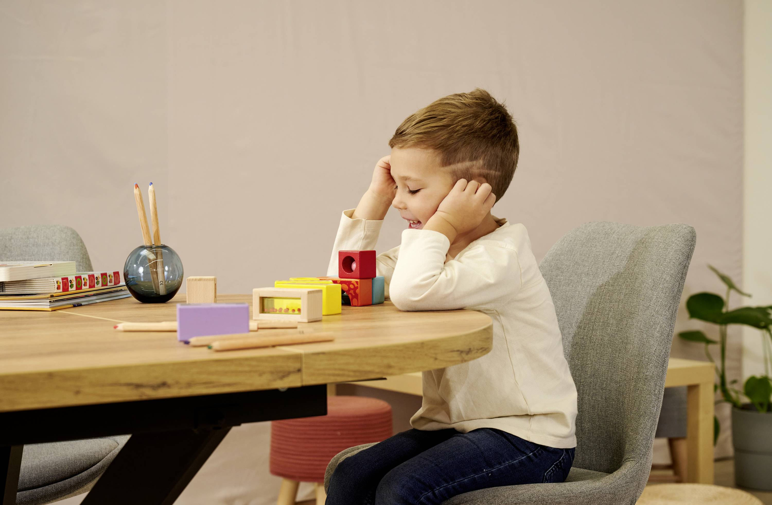 Ein Kind sitzt entspannt am Tisch und spielt mit bunten Holzklötzen. Im Hintergrund sind Stifte in einem Behälter aufgestellt.